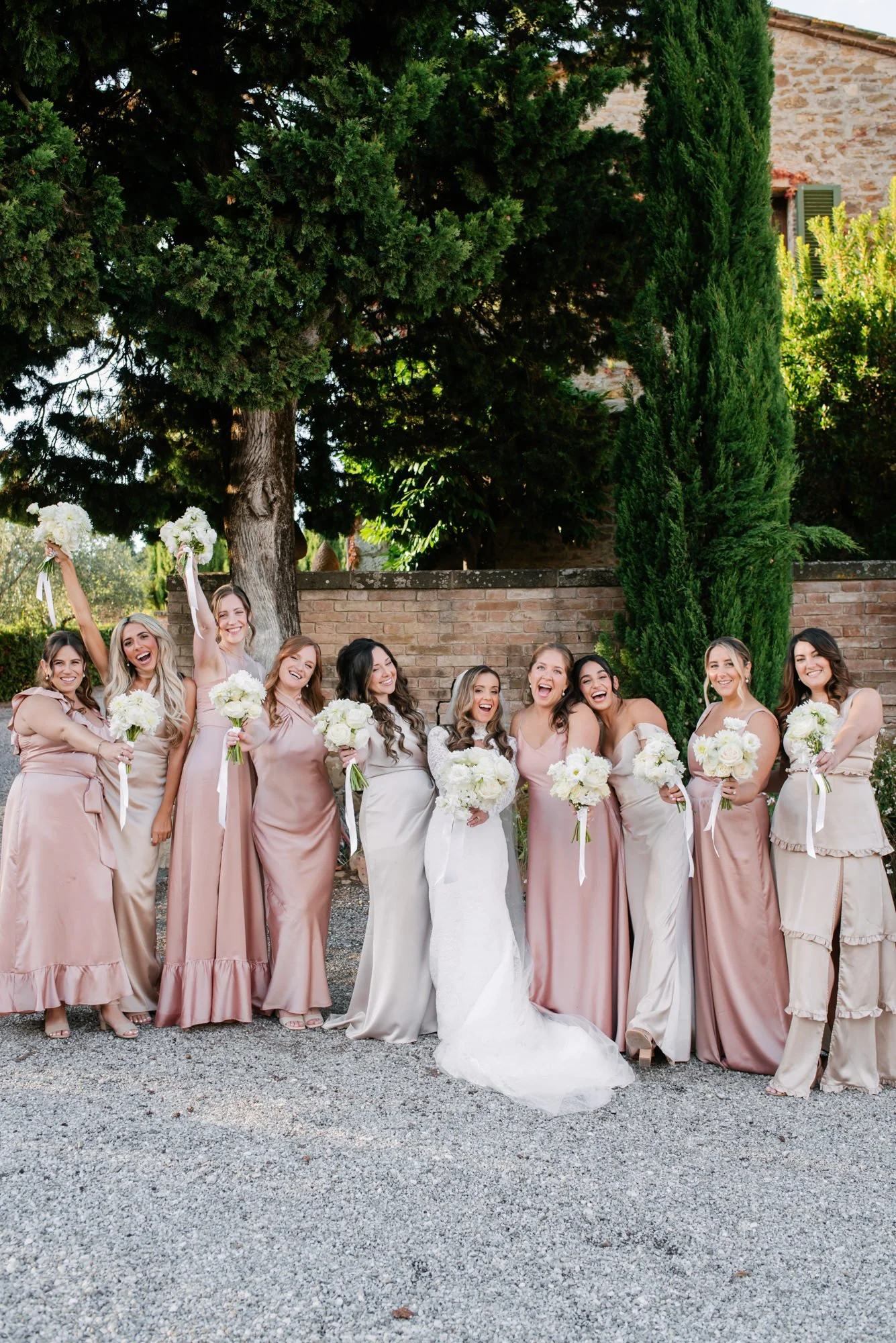 Bride and bridesmaids smiling and celebrating outdoors, holding white bouquets, standing on gravel ground near a brick wall and trees.