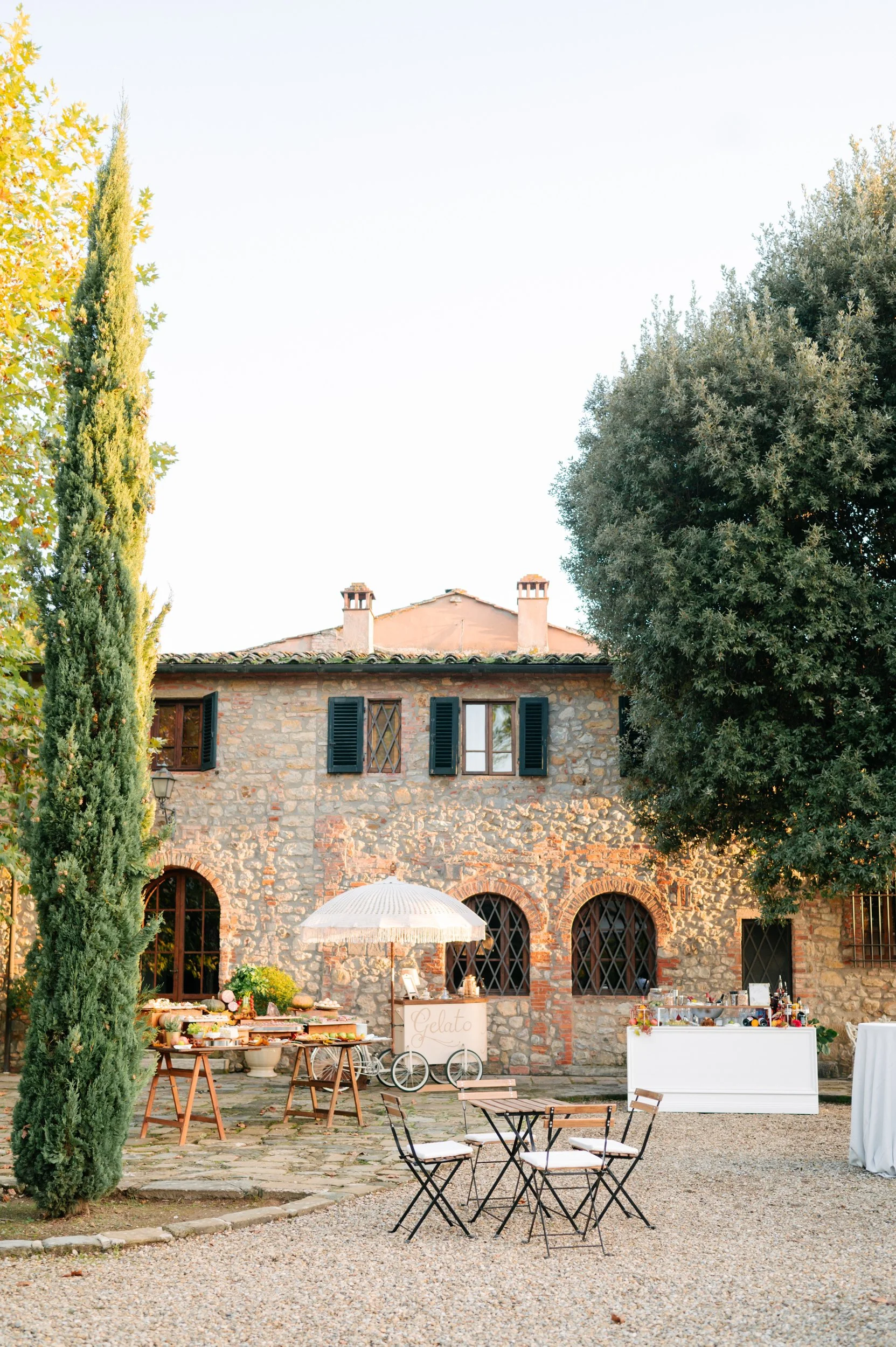 Outdoor wedding or event setup in front of a rustic stone house with black window shutters, surrounded by trees, with tables and chairs, a gelato cart with umbrella, and buffet stands.