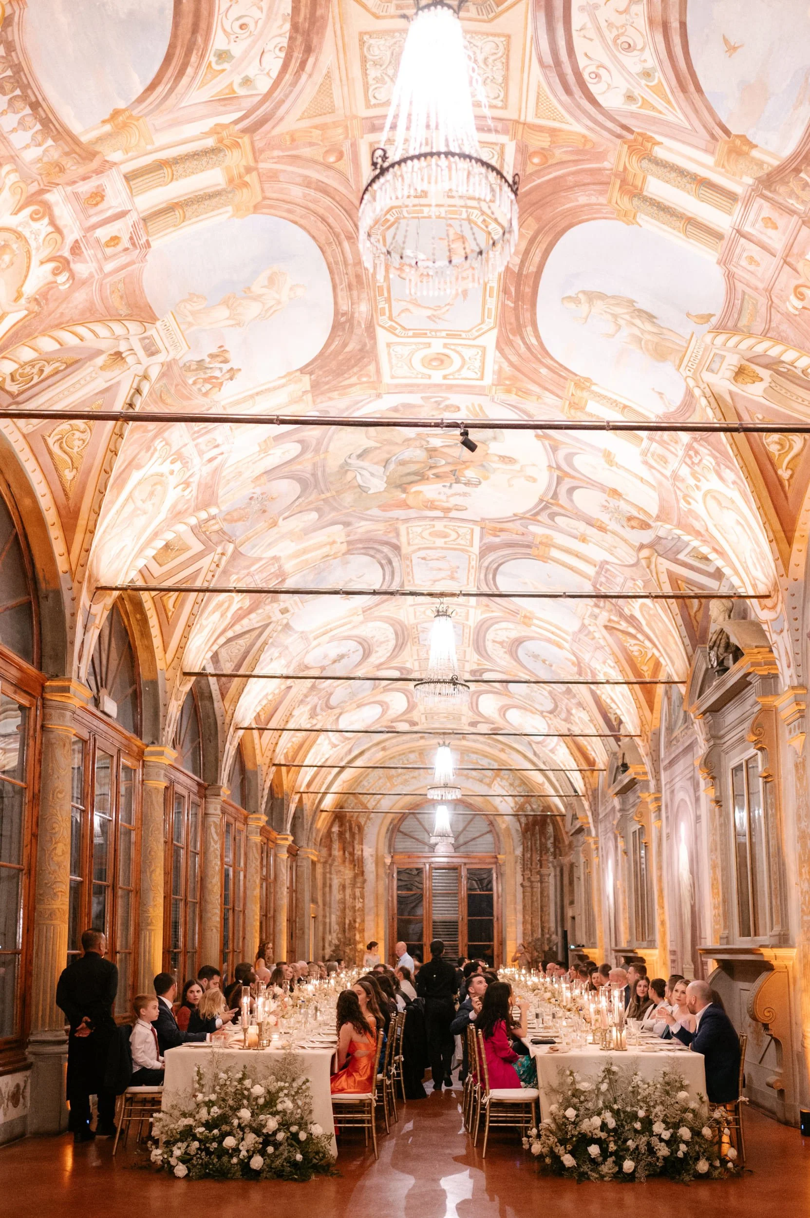 Elegant banquet hall with a high, ornate painted ceiling, chandeliers, and large windows. Long dining tables are set with white tablecloths, candles, and floral arrangements, with guests seated on gold chairs.