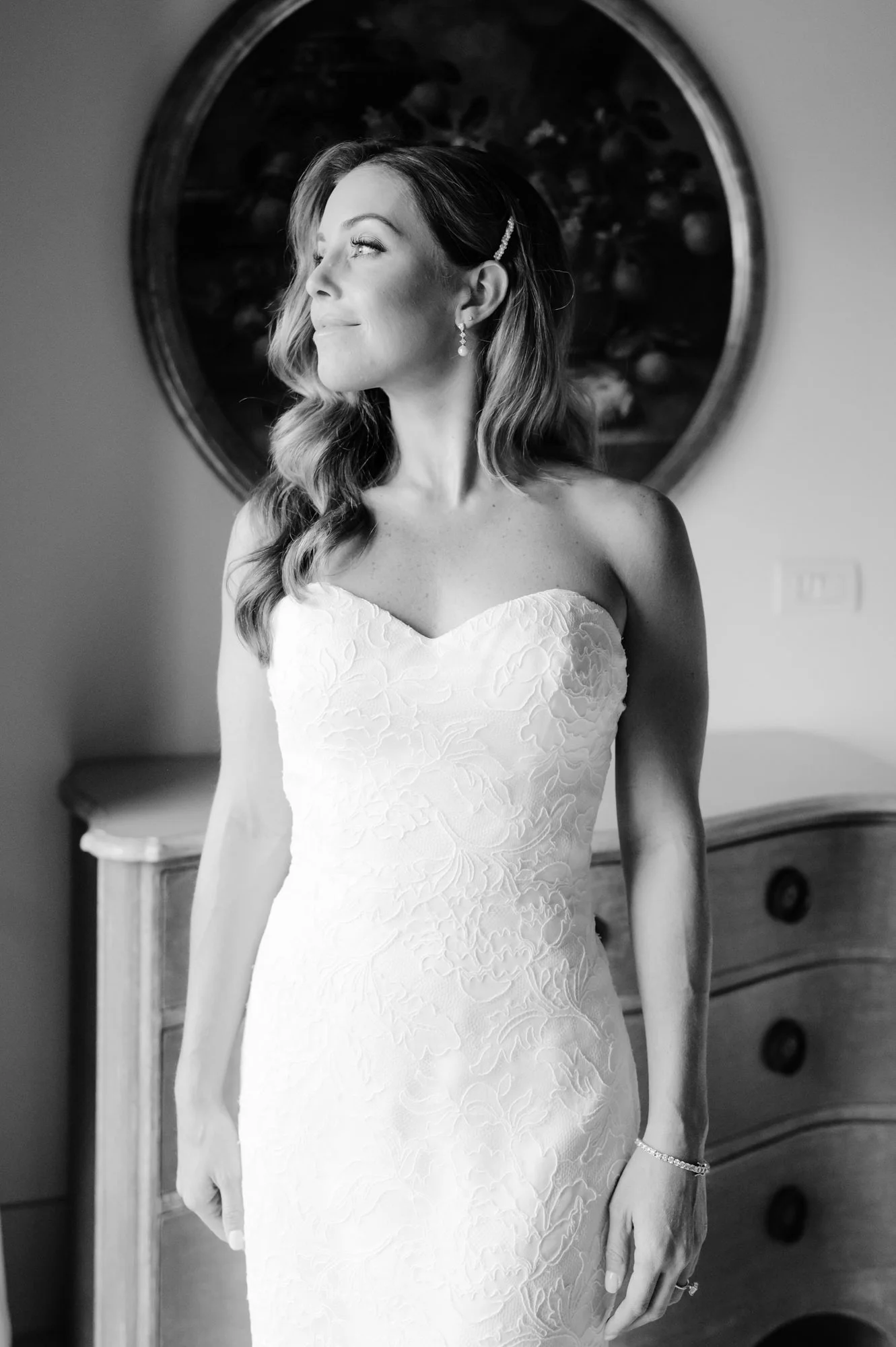 A bride in a strapless wedding dress with a floral pattern, standing indoors in front of a mirror. She has wavy hair, earrings, and a bracelet, looking off to the side with a gentle smile. Luxury editorial wedding photography in Tuscany. Destination 