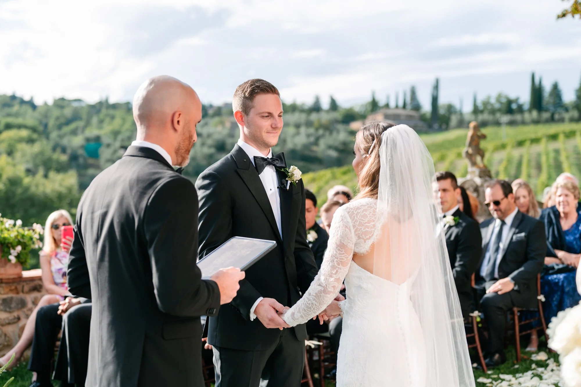 A couple gets married outdoors on a sunny day, with the groom in a black tuxedo and the bride in a lace white wedding dress and veil, exchanging vows while holding hands. The officiant stands nearby with a book, and guests are seated behind them, wit