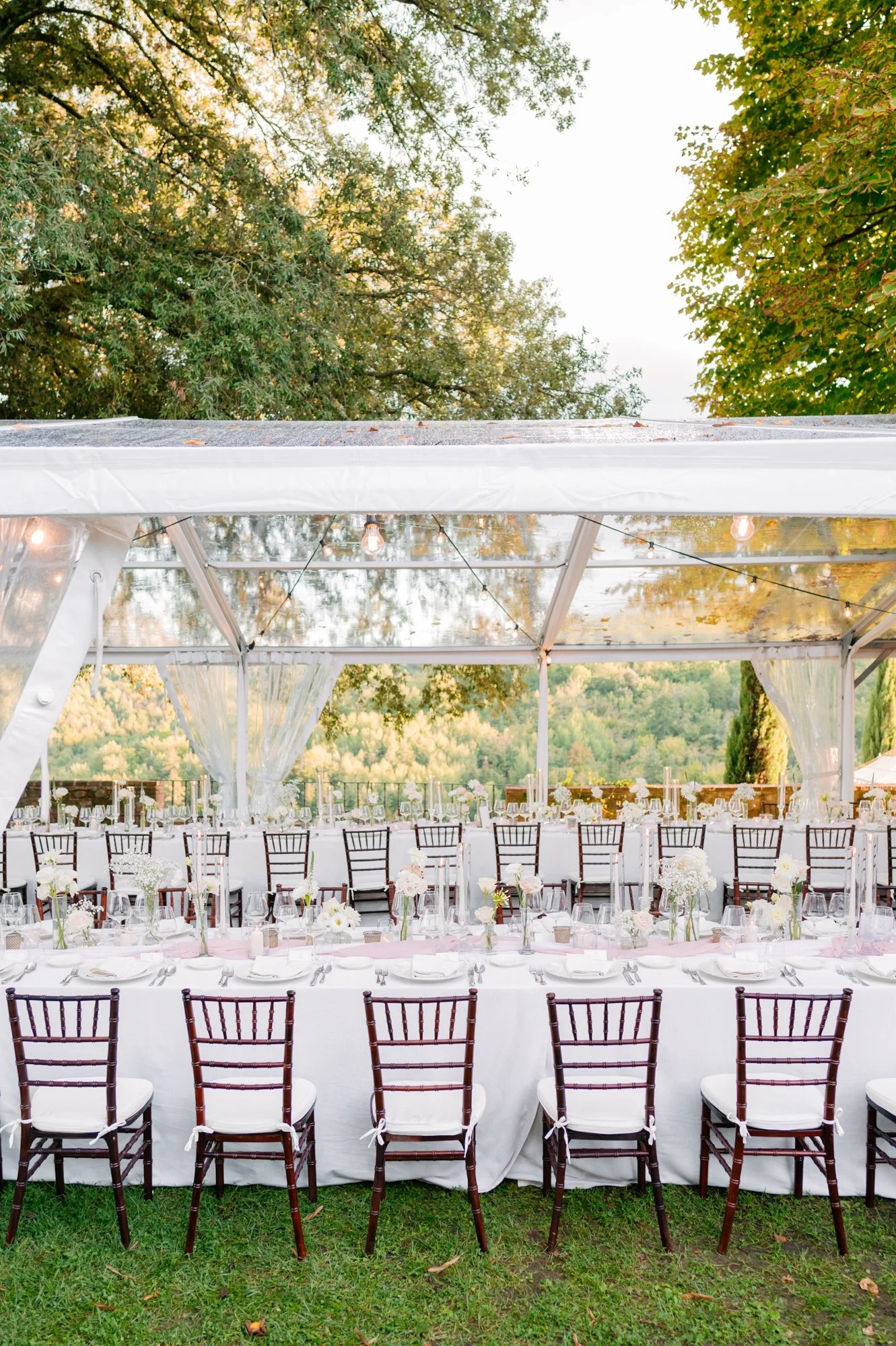 Outdoor event space with a large white tent, decorated with string lights, and surrounded by greenery.