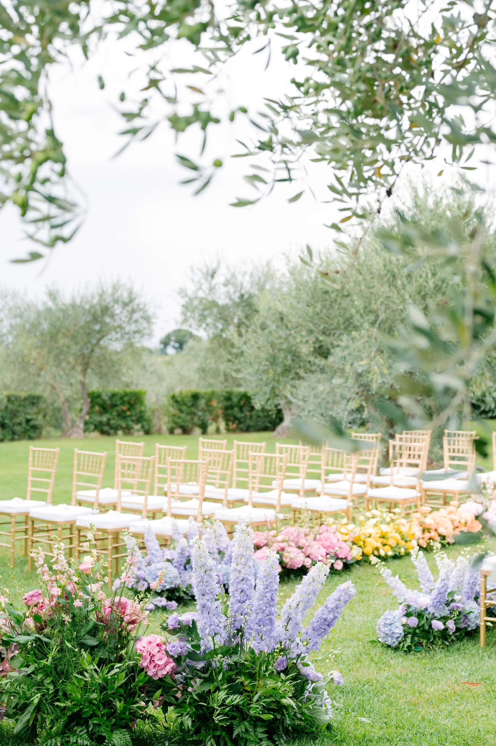 Outdoor wedding ceremony setup with rows of gold chairs with white cushions and colorful floral arrangements on a green lawn, surrounded by trees.