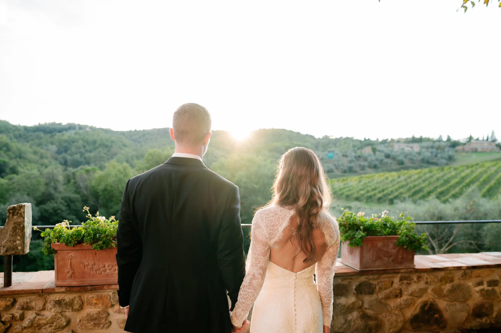 A newlywed couple holding hands on a balcony overlooking a lush green landscape at sunset.