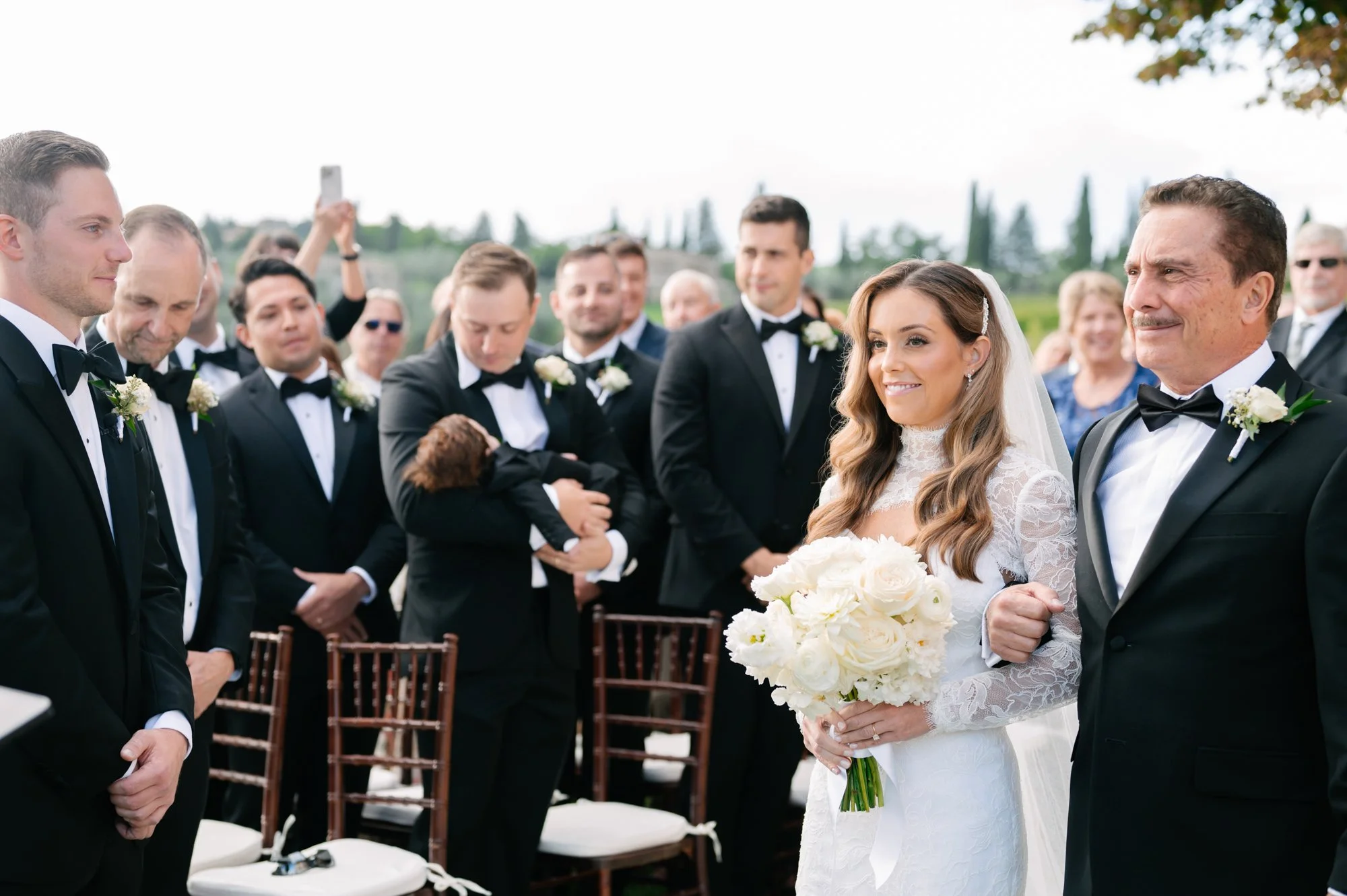 Bride and father walking down the aisle at an outdoor wedding, surrounded by groomsmen and guests in formal attire, with greenery and trees in the background.