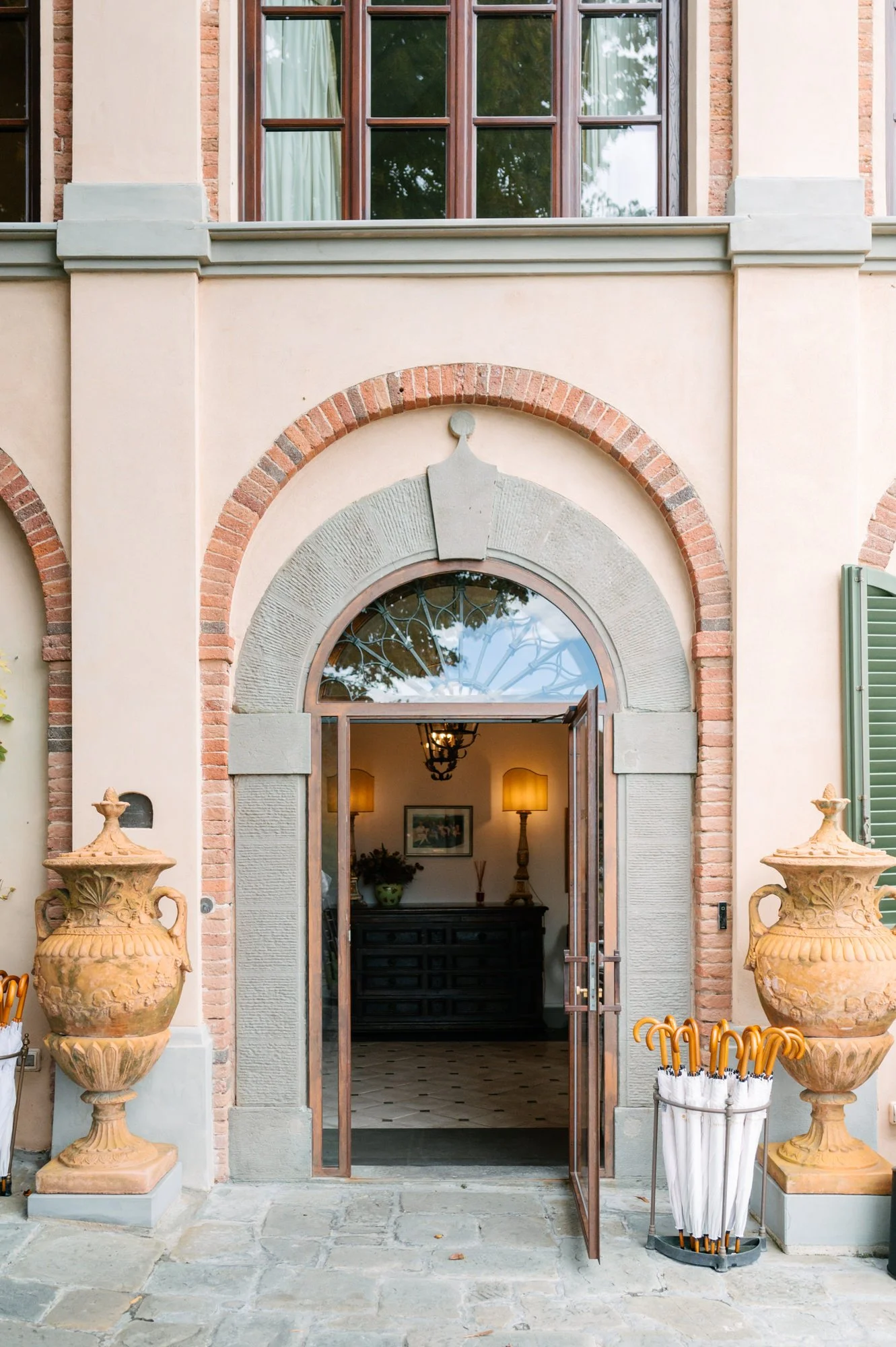 The entrance of a building with an open door, flanked by two large decorative urns, and an umbrella stand with umbrellas outside.