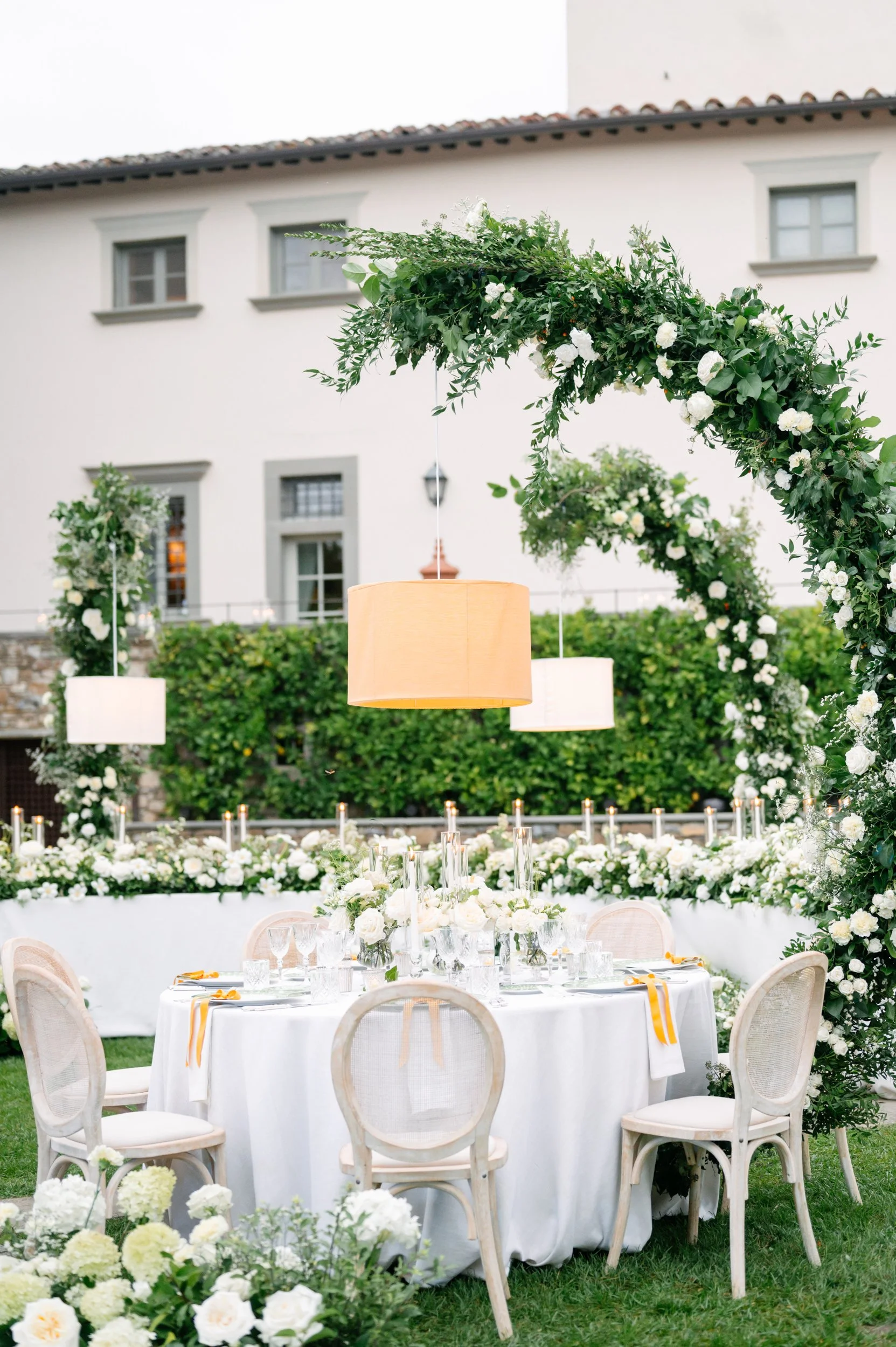 Outdoor wedding reception setup with round table, white tablecloth, floral centerpieces, surrounded by chairs, decorated arch with white flowers and greenery, hanging lamps, and a lush garden background.
