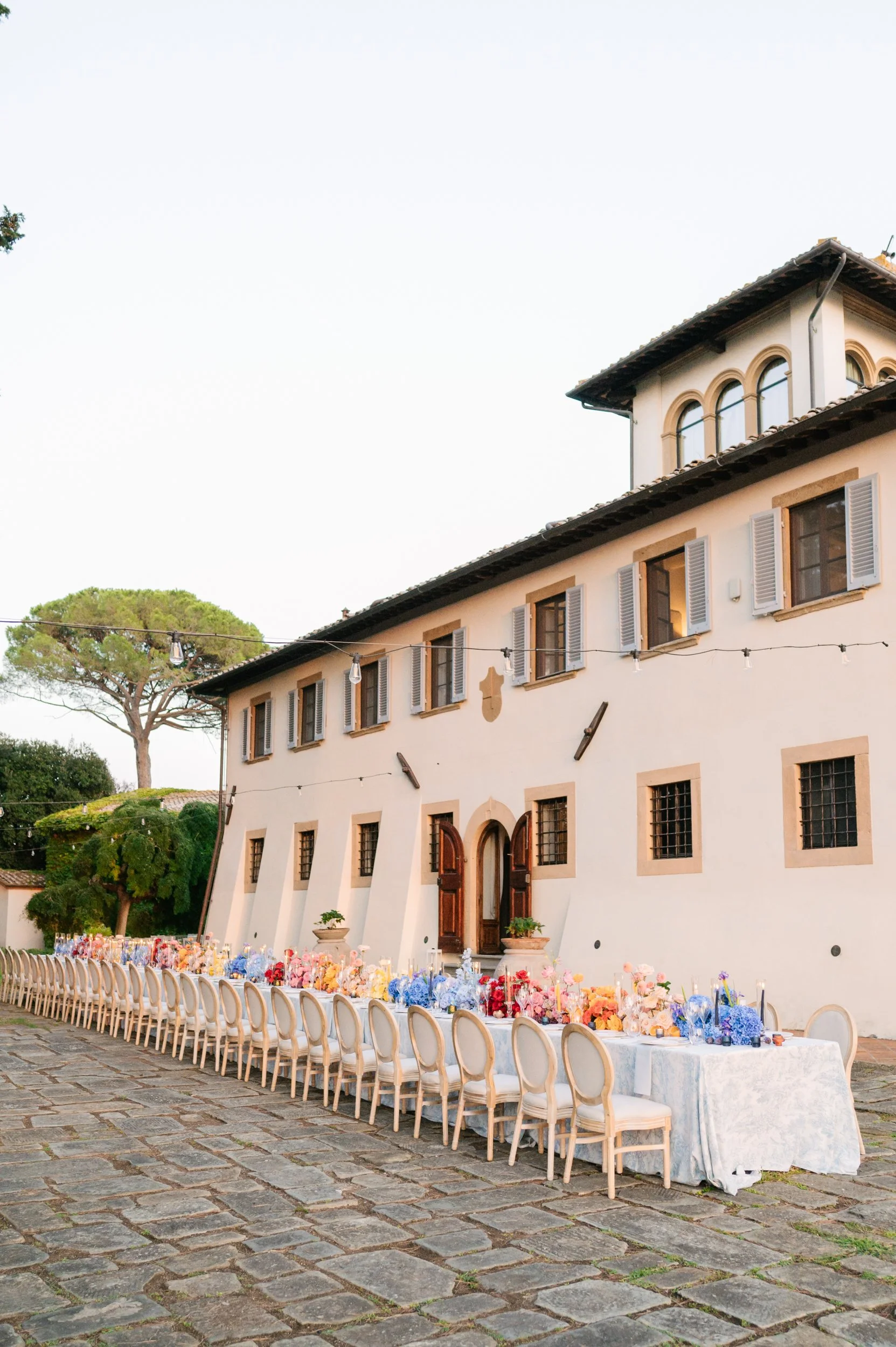Long dining table with chairs and floral arrangements outside a white building
