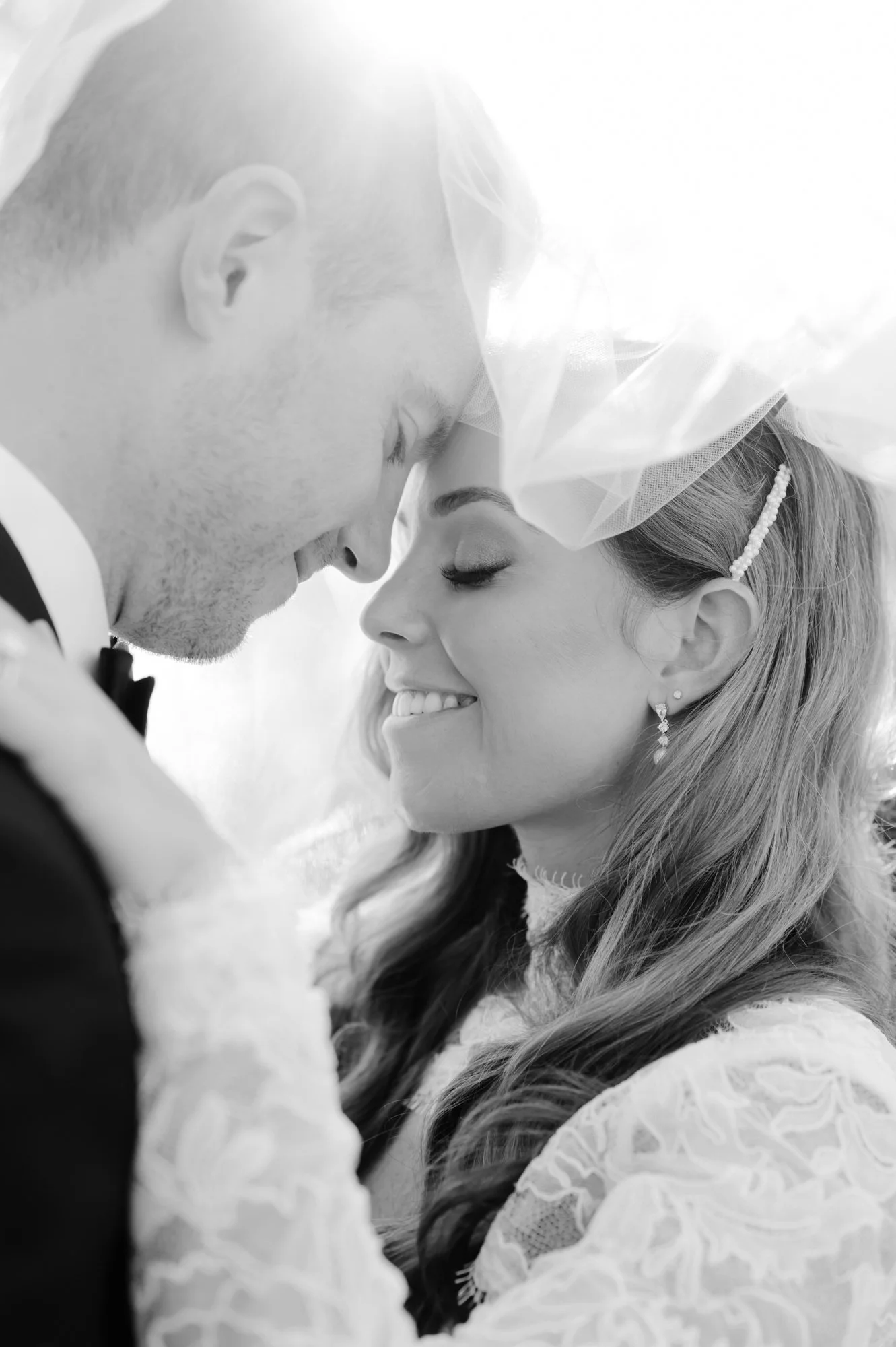 A black and white close-up photo of a bride and groom with their foreheads touching, smiling with eyes closed, on their wedding day.