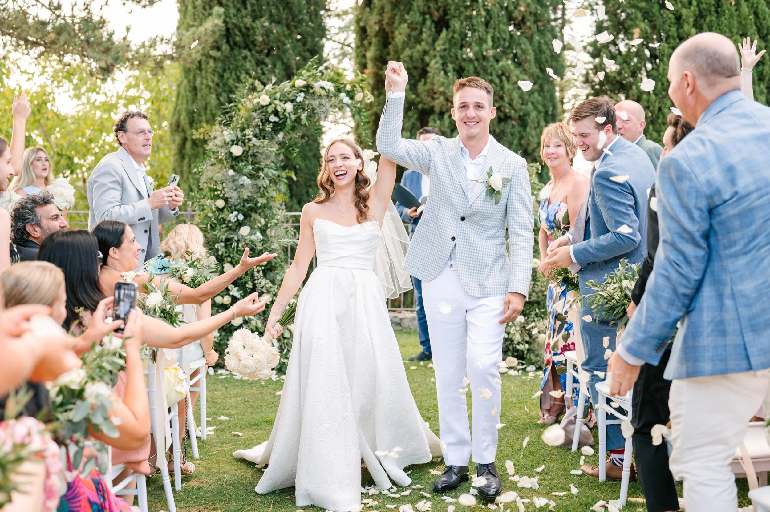 A newlywed couple is walking down the aisle holding hands, smiling, while guests celebrate and throw flower petals around them at an outdoor wedding ceremony under trees.