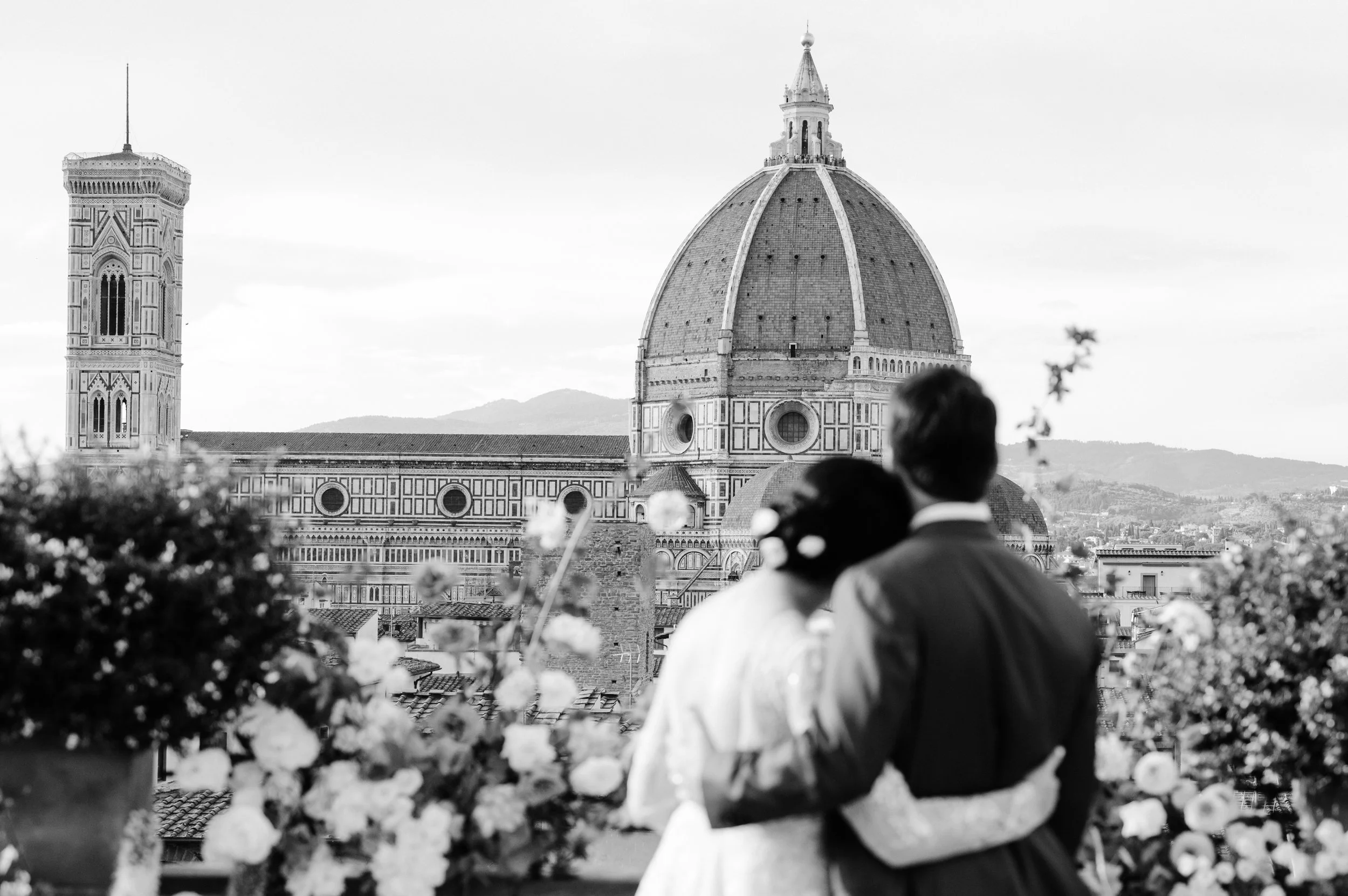 A couple, possibly newlyweds, with their backs to the camera, embrace on a rooftop terrace adorned with flowers, with Florence Cathedral's dome and cityscape in the background.