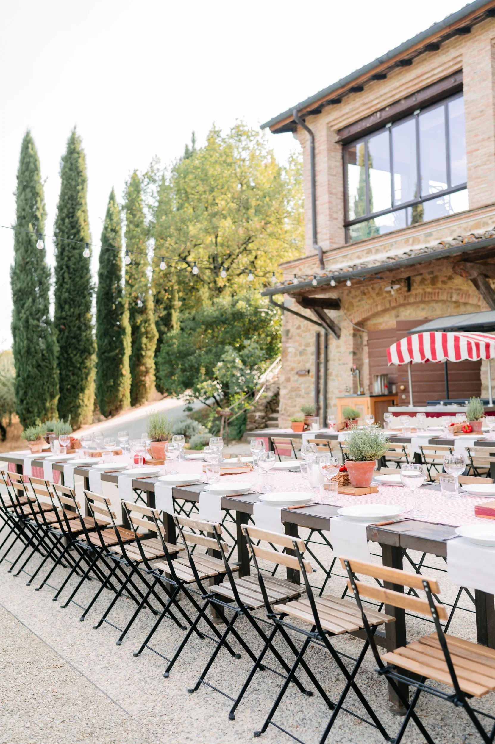 Outdoor dining setup with a long table, chairs, tableware, potted plants, and a brick house with large windows, set in a garden with tall trees and string lights.