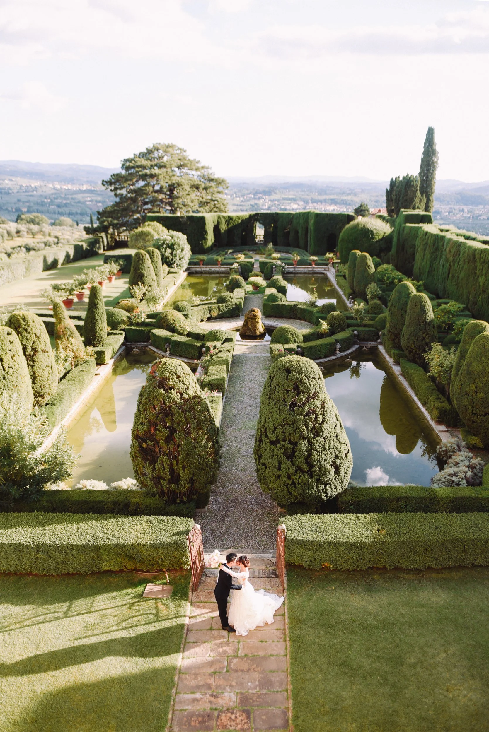 A bride and groom dancing at the entrance of a beautifully landscaped garden with trimmed hedges, water features, and scenic distant hills in the background.