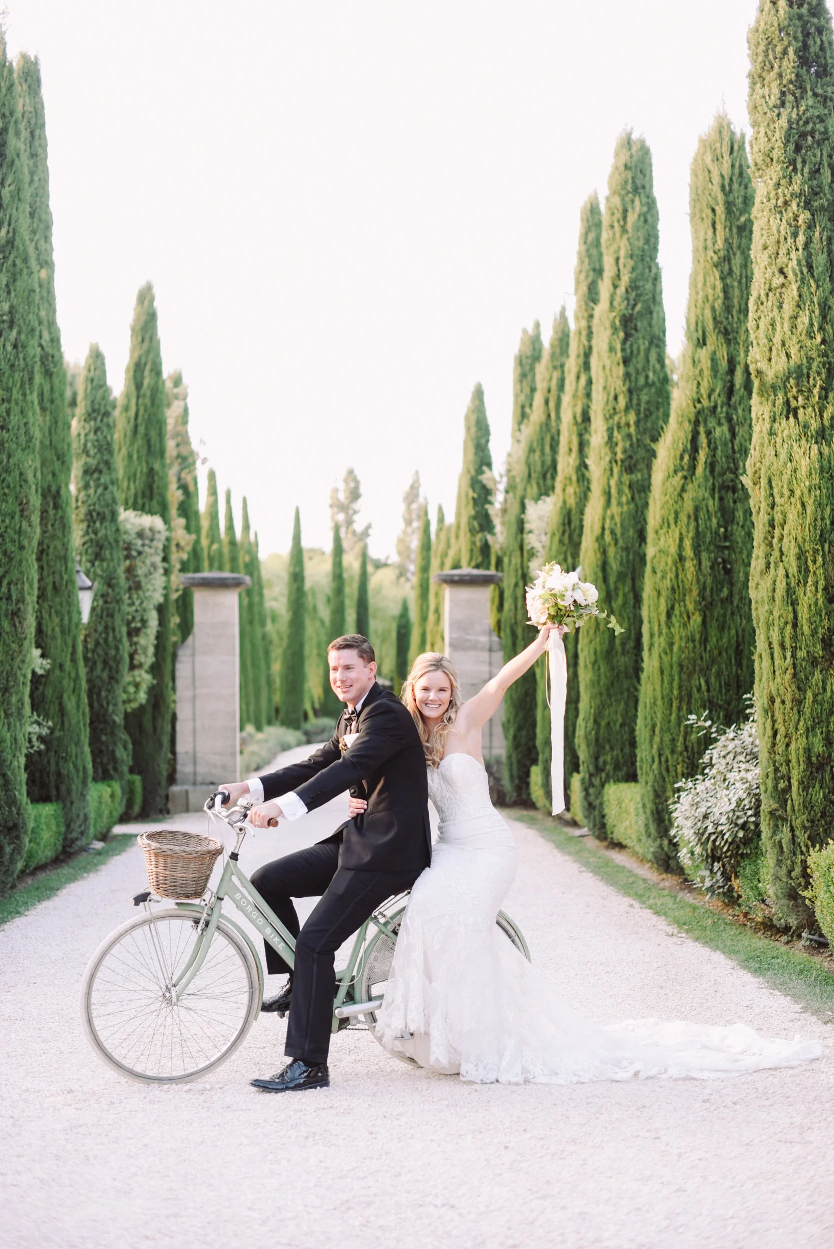 A bride and groom celebrating their wedding ride on a bicycle through a lush, green pathway lined with tall, trimmed trees. The bride, in a white lace wedding gown, is sitting on the back holding a bouquet of white flowers in the air, while the groom