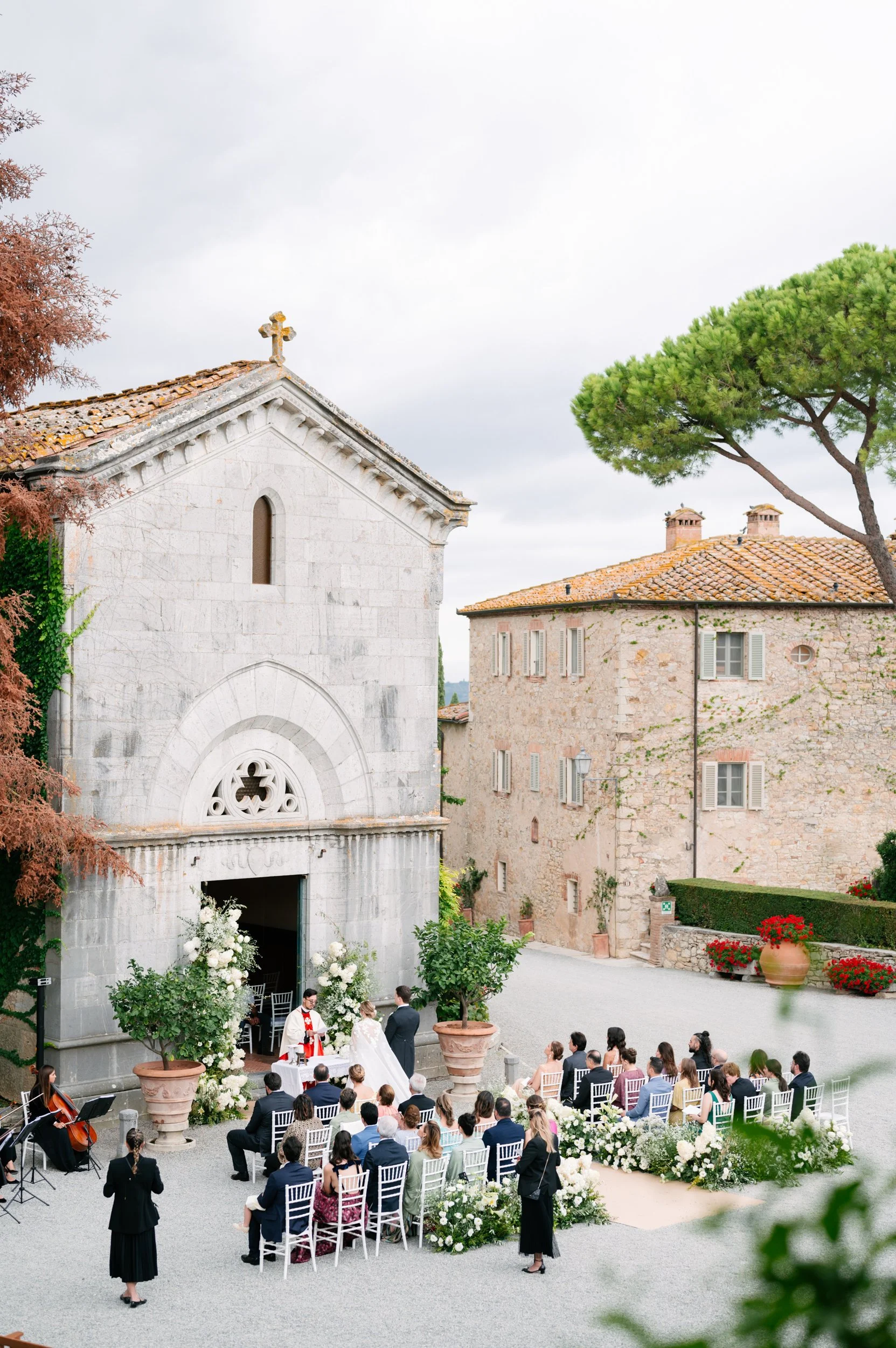 A wedding ceremony taking place outdoors near a historic stone church, with guests seated on white chairs, a bride and groom at the altar, and musicians playing nearby, surrounded by trees and flowers.