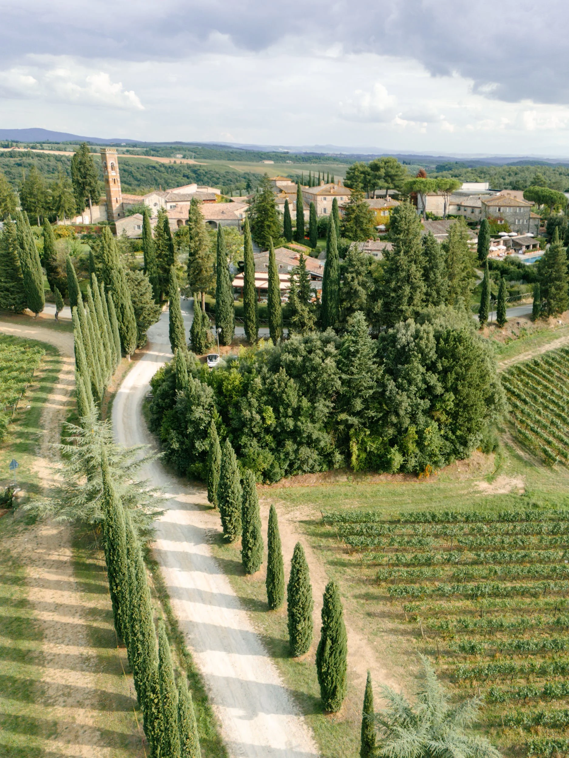 Aerial view of a small European village with a church, surrounded by cypress trees, vineyards, and rolling hills.