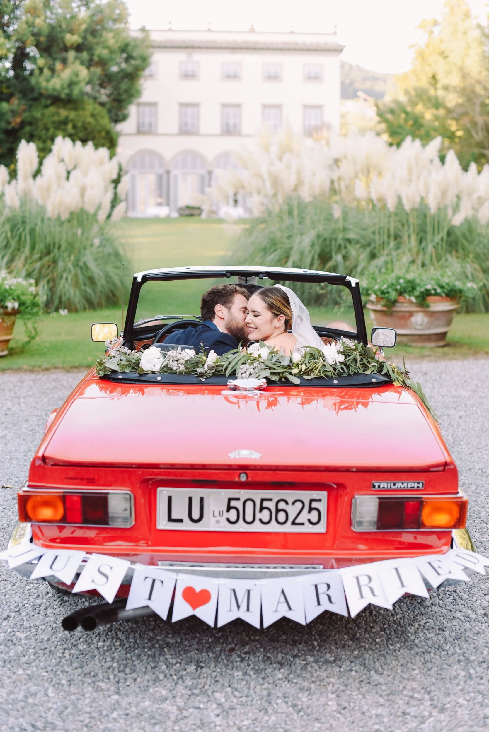 A newlywed couple in a red Triumph convertible decorated with flowers and a 'Just Married' banner, sharing a kiss before a vintage-style house with lush greenery.