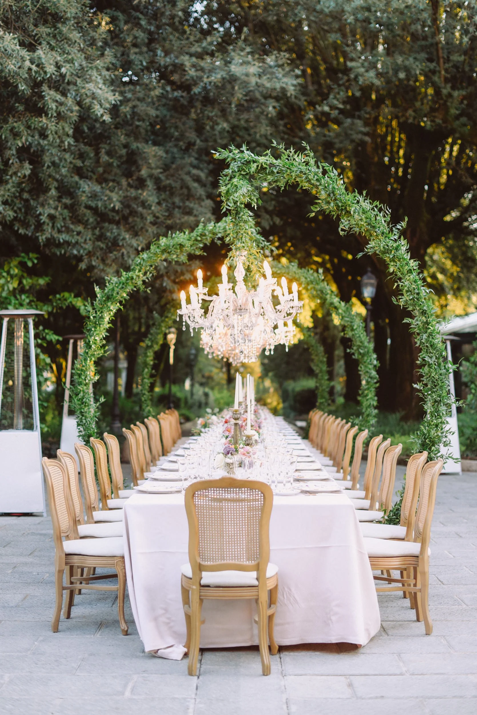 Long outdoor dining table set with white tablecloths, elegant glassware, and floral centerpieces, under a crystal chandelier and green leafy arches, surrounded by trees.
