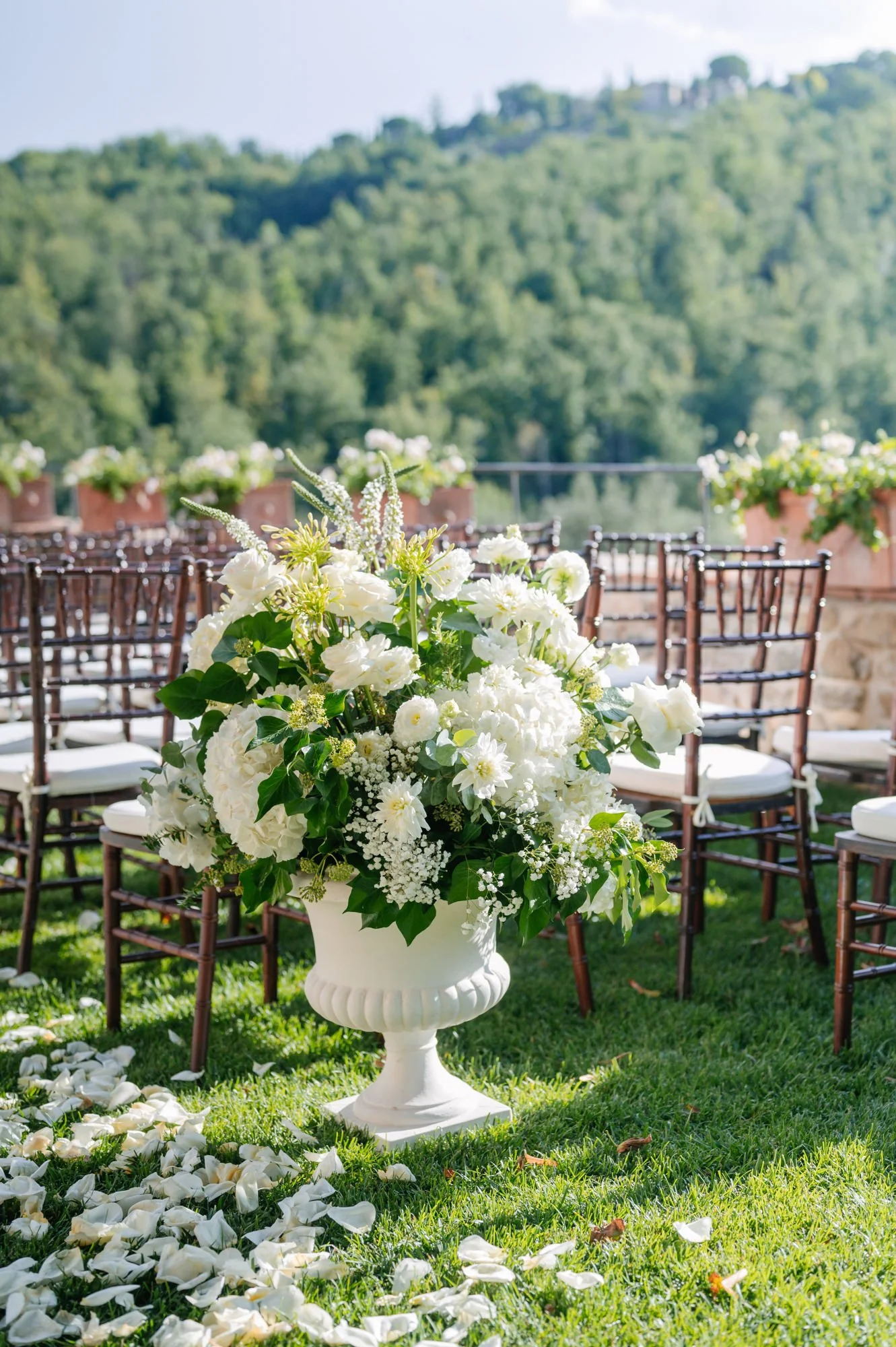 A white wedding flower arrangement in a vase, placed in front of wooden chairs with white cushions, on a grassy outdoor setting with hills and trees in the background.