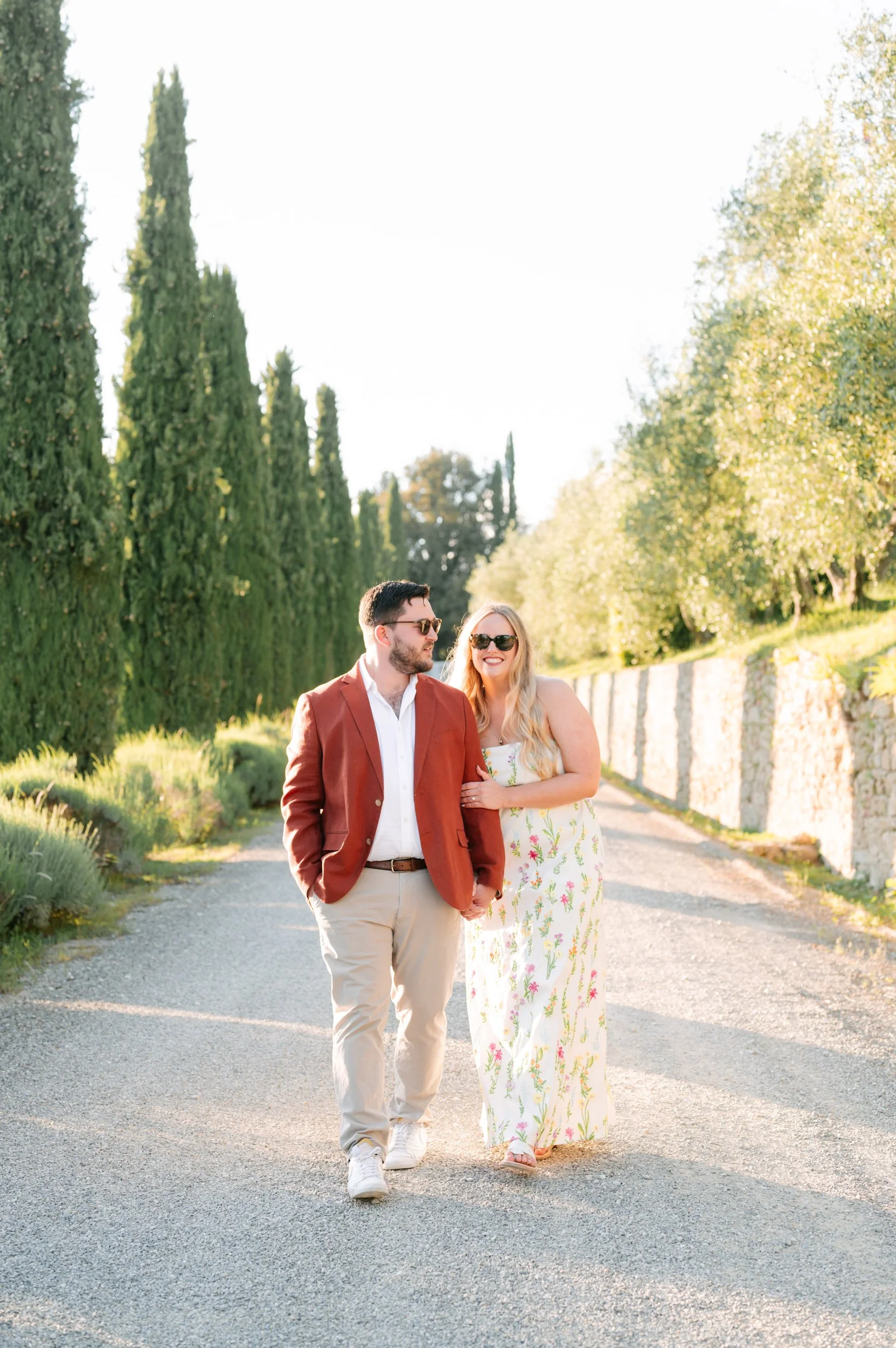 A man and woman walk together on a gravel path surrounded by green trees and plants during the daytime. They are both wearing sunglasses, and the woman is smiling and has her arm linked with the man.