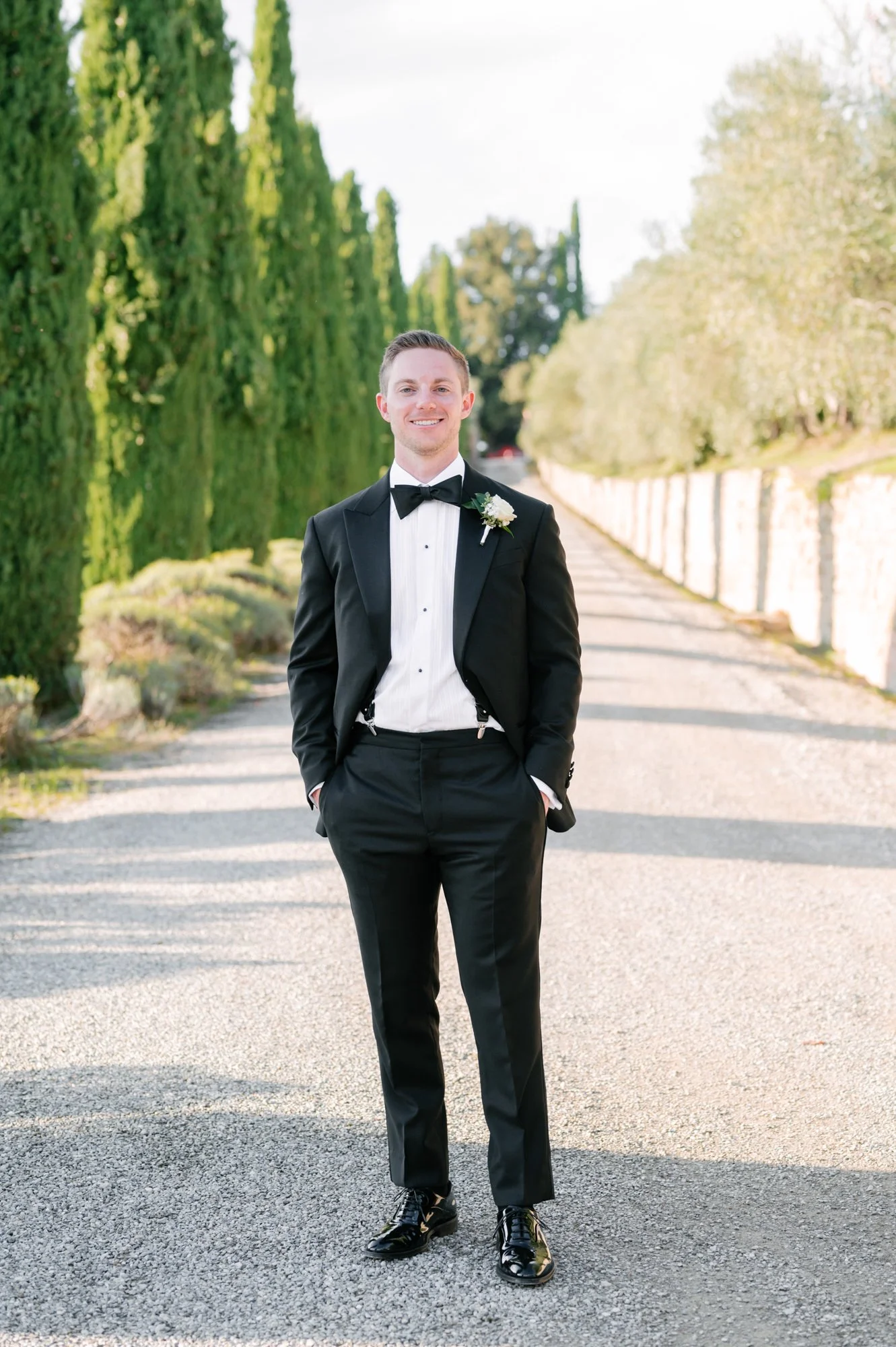 Man dressed in a black tuxedo with a bow tie, standing outdoors on a paved pathway with greenery and trees in the background, smiling at the camera.
