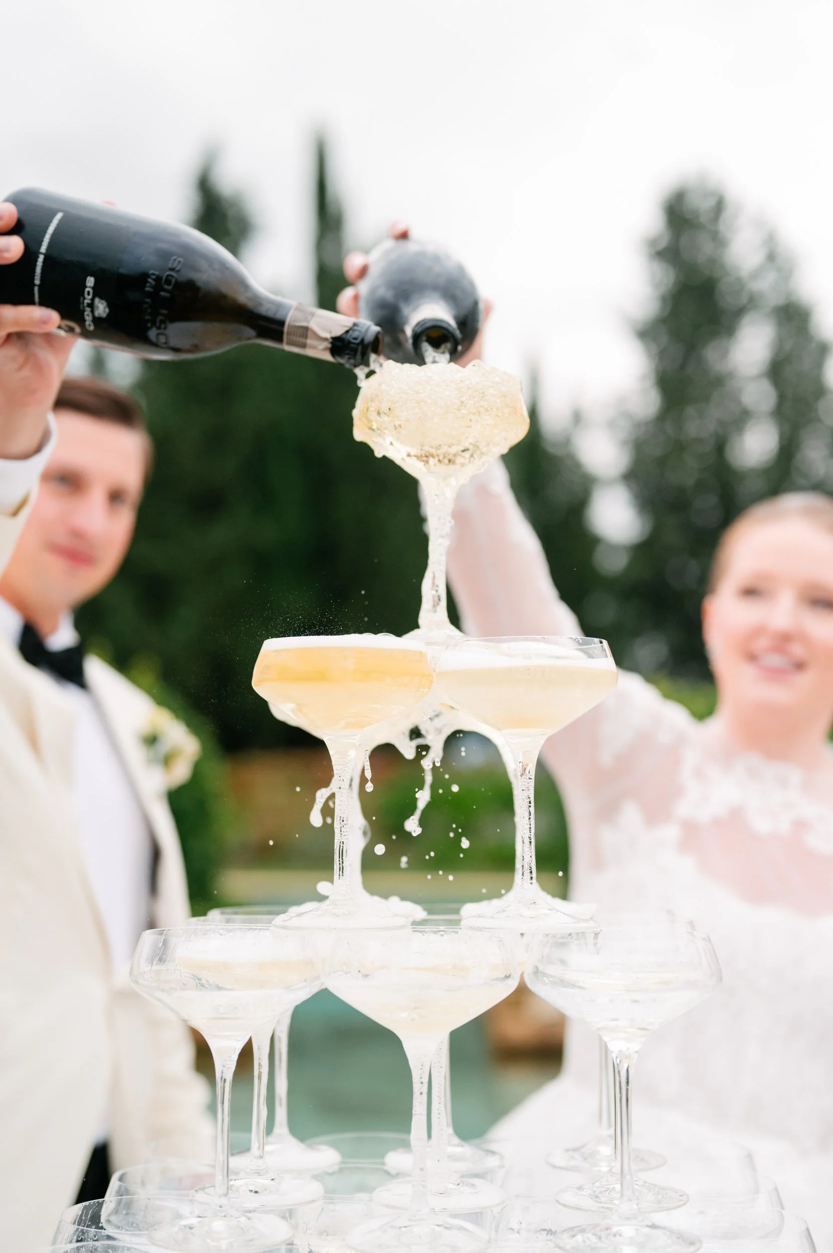 People celebrating outdoors by pouring champagne into a champagne tower at a wedding reception.