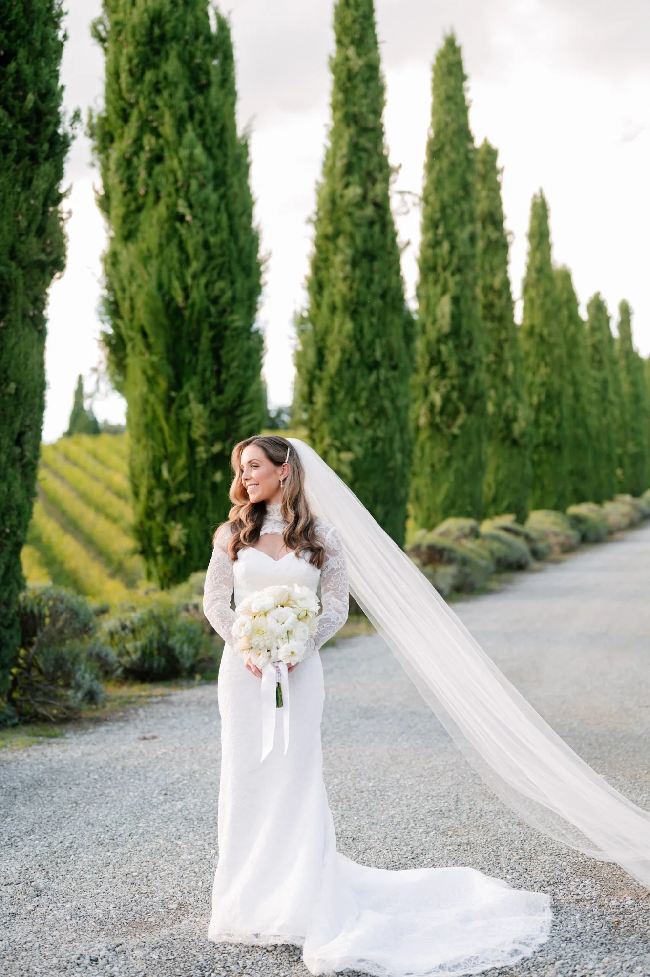 A smiling bride in a white wedding dress with lace sleeves, holding a bouquet of white flowers, standing on a gravel path lined with tall green cypress trees during daytime.