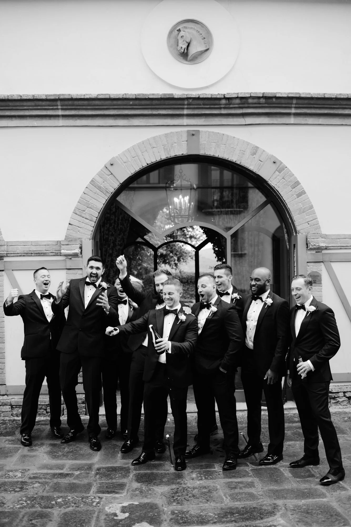 A group of men dressed in tuxedos celebrating outside a building with an arched window and a circular emblem with a horse head above. Luxury editorial wedding photography in Tuscany. Destination wedding in Italy. L&V Photography Letizia Maccarini.