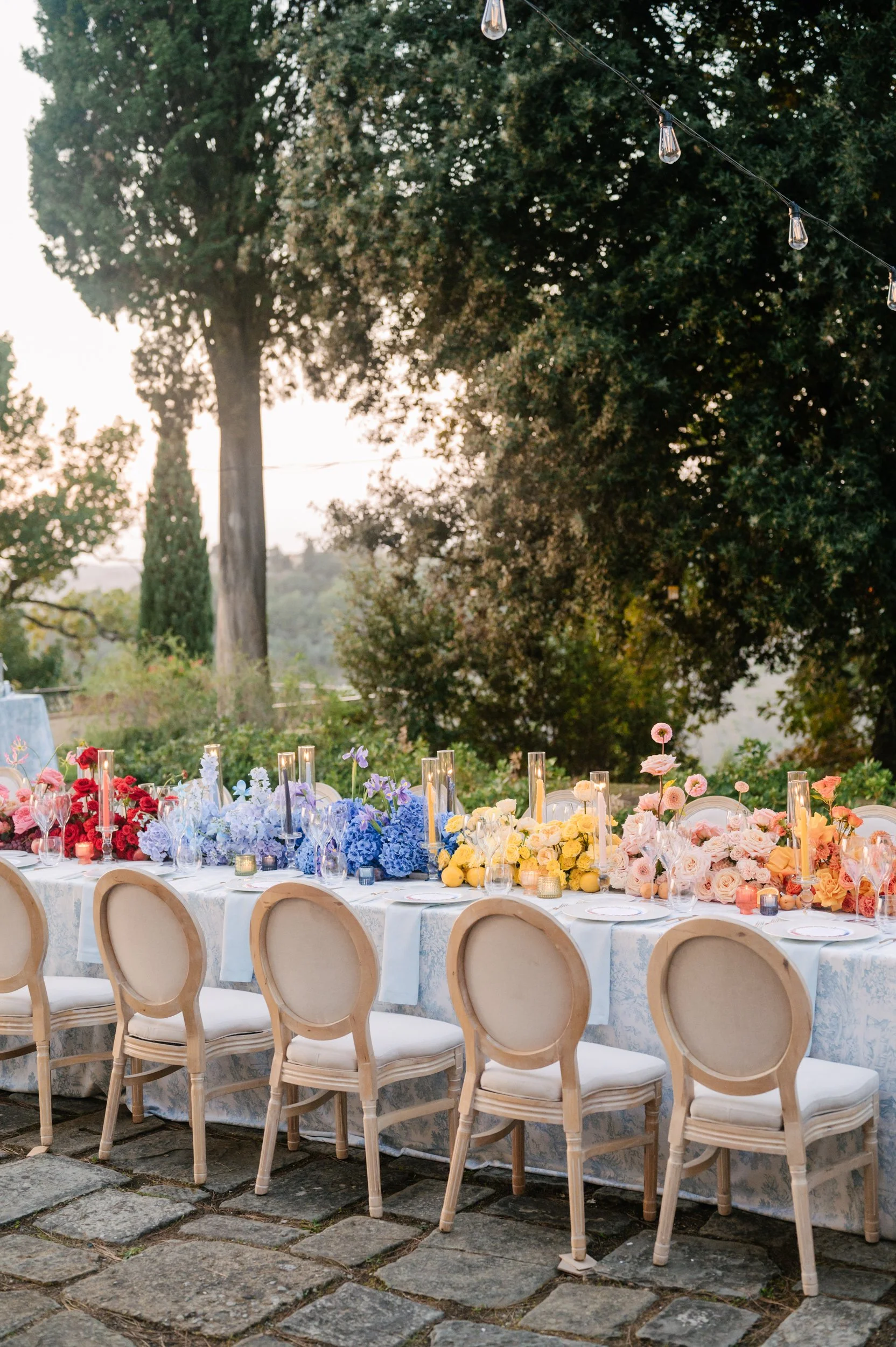 A long banquet table outdoors decorated with colorful flowers and candles, set for a celebration or event, with chairs on one side and string lights overhead, surrounded by trees.