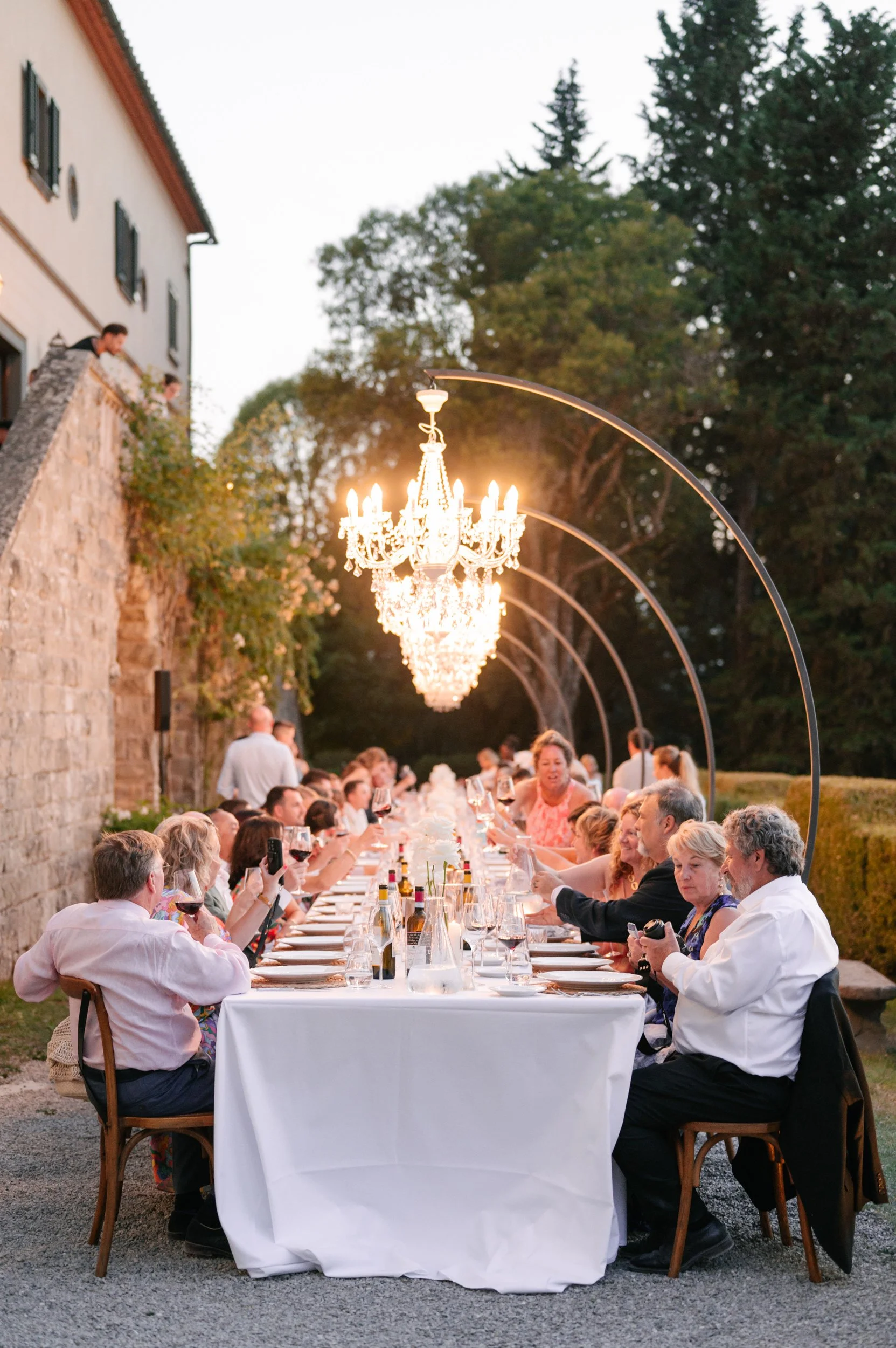 An outdoor dinner party with a long table, chandeliers overhead, and guests enjoying food and drinks during dusk.