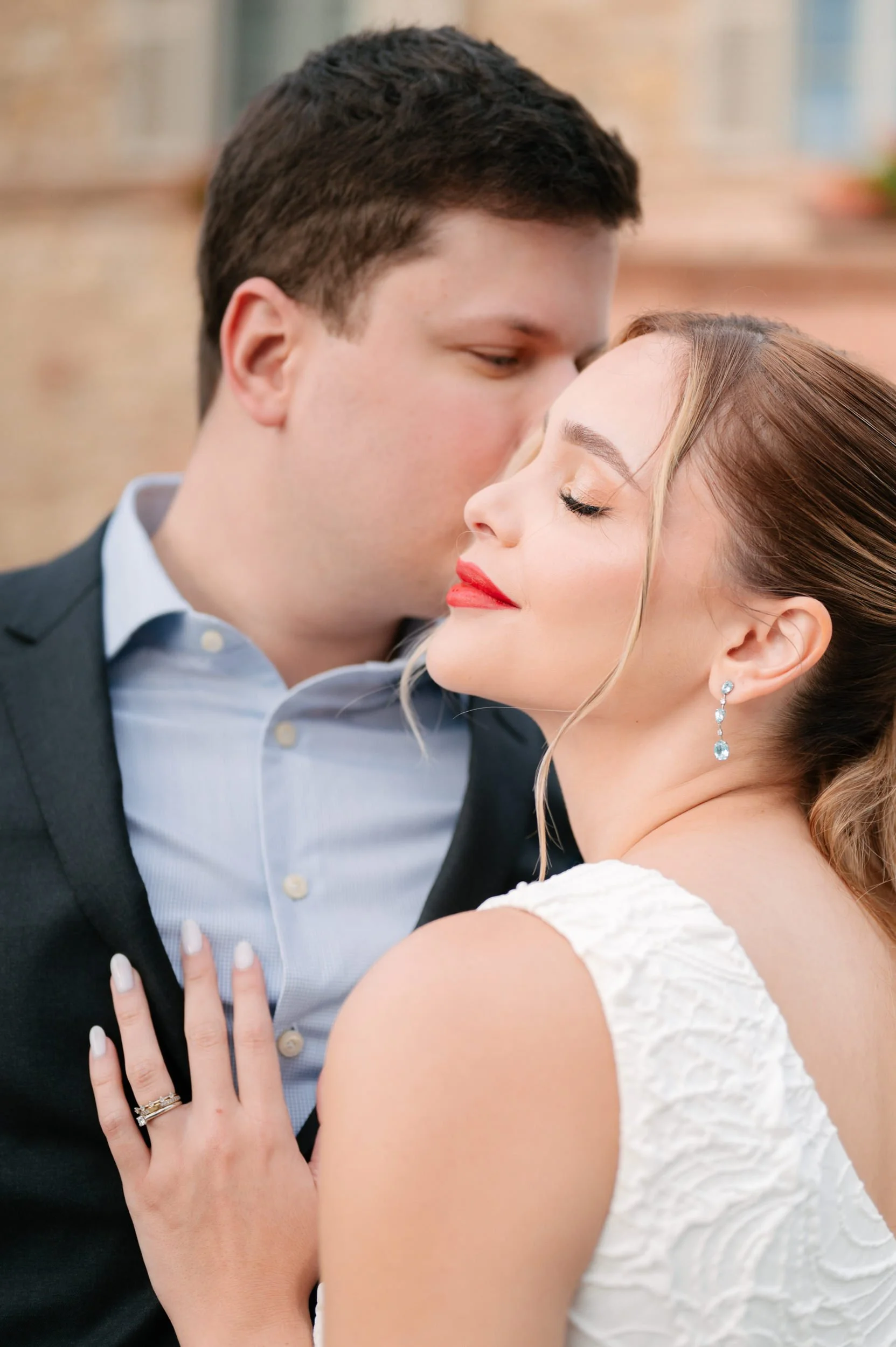 A couple in formal attire sharing a close moment, with the man gently kissing the woman's forehead. The woman has her eyes closed, wears bright red lipstick and elegant earrings, and has her hand resting on the man's chest showing a ring.