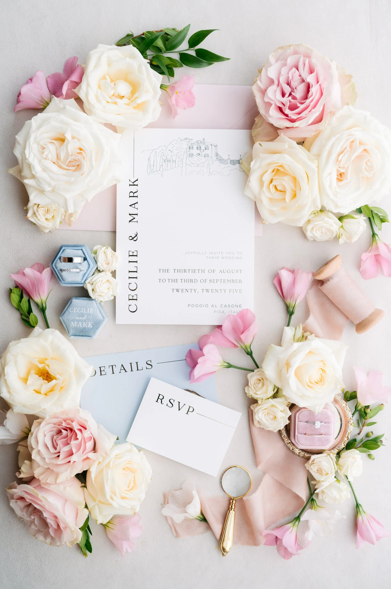 Wedding invitation surrounded by white and pink flowers, rings in a blue velvet box, pink velvet ring box, and pink fabric.