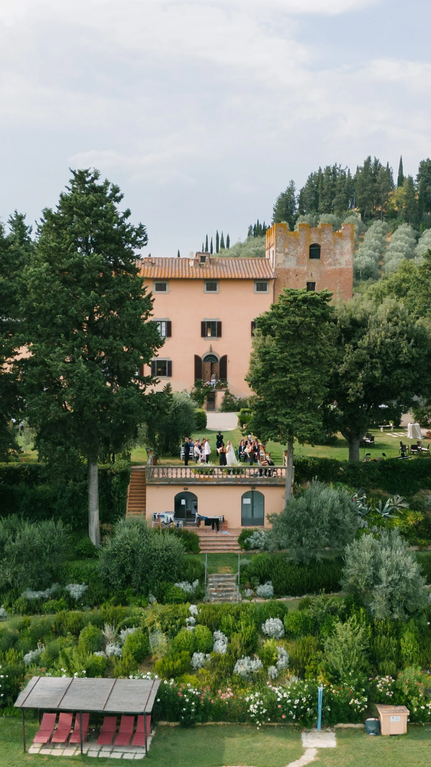 A large pink villa with a tower, surrounded by green trees and a well-maintained garden. There is a group of people dressed formally gathered on the lawn, indicating a celebration or event.