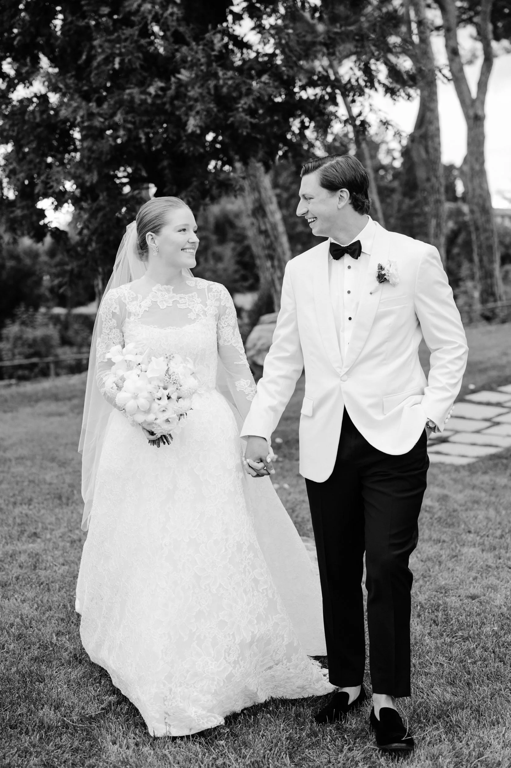 Black and white photo of a bride and groom walking hand in hand outdoors, smiling at each other. The bride is in a lace wedding gown holding a bouquet, and the groom is in a white tuxedo jacket with a black bow tie.