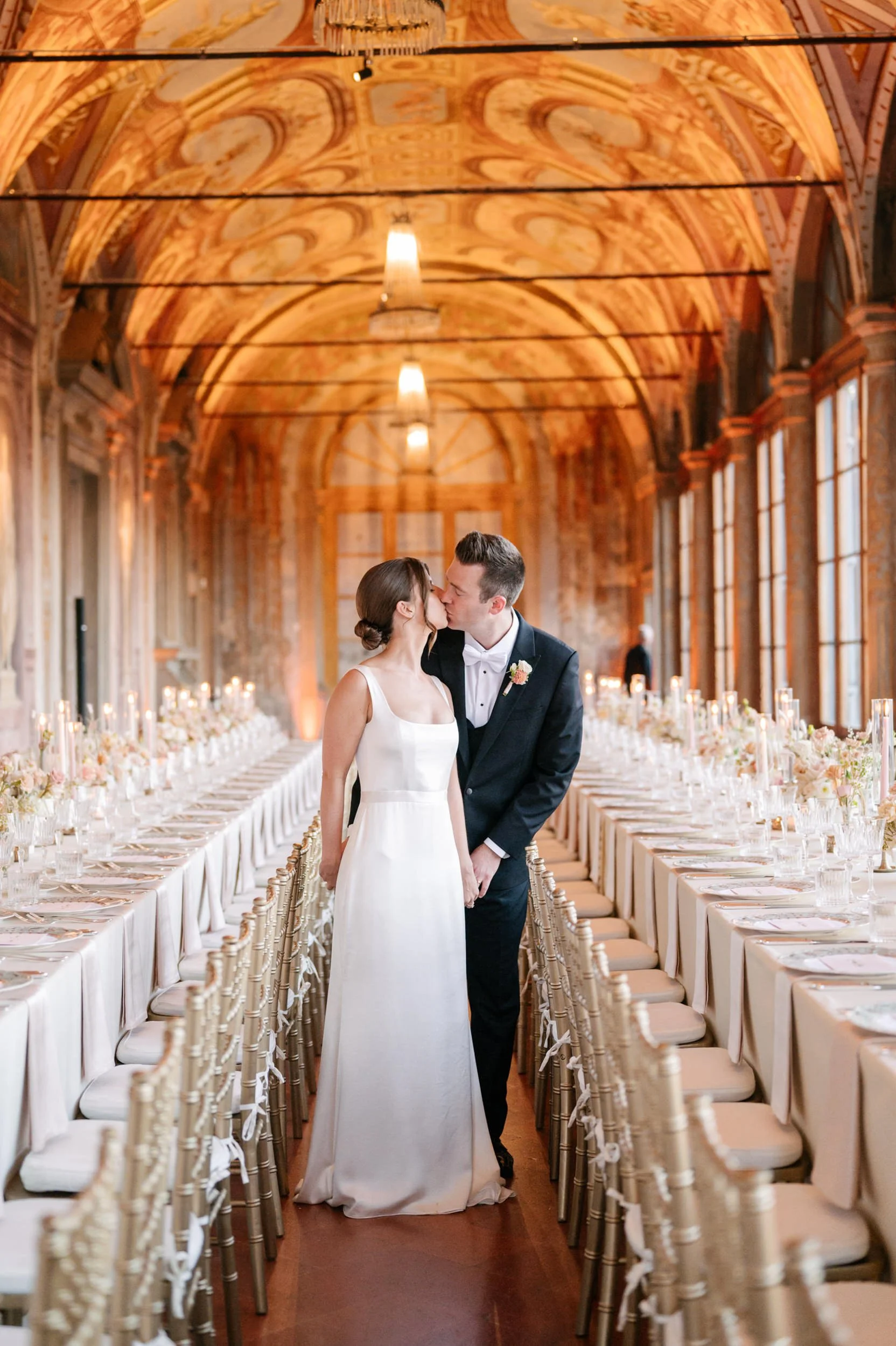 A bride and groom sharing a kiss in a decorated banquet hall with long tables, candles, floral centerpieces, and ornate architectural details.