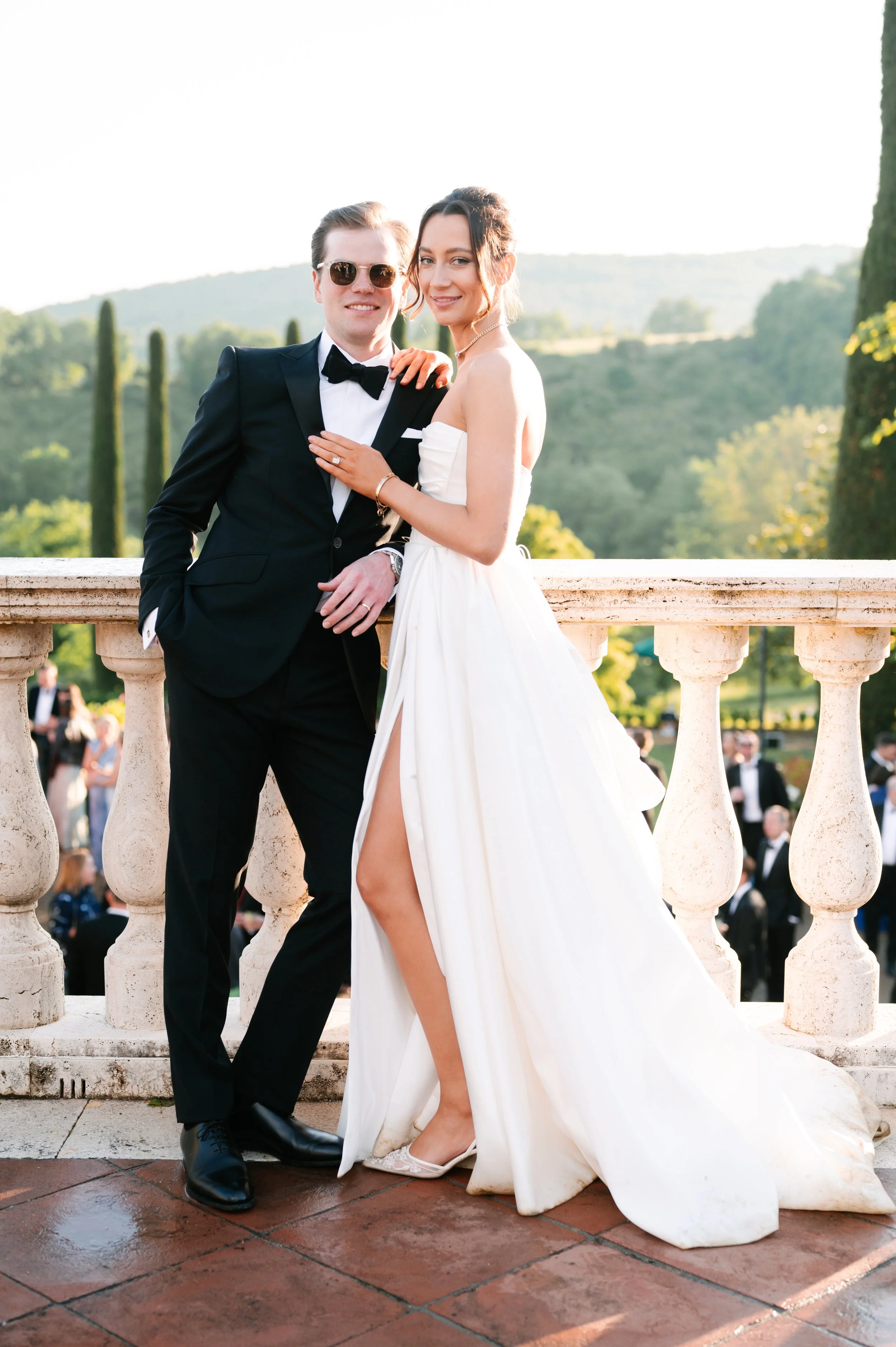 A newlywed couple in wedding attire standing on a balcony with scenic greenery and rolling hills in the background. The groom wears a black tuxedo with a bow tie, and the bride wears a strapless white wedding gown with a high slit.