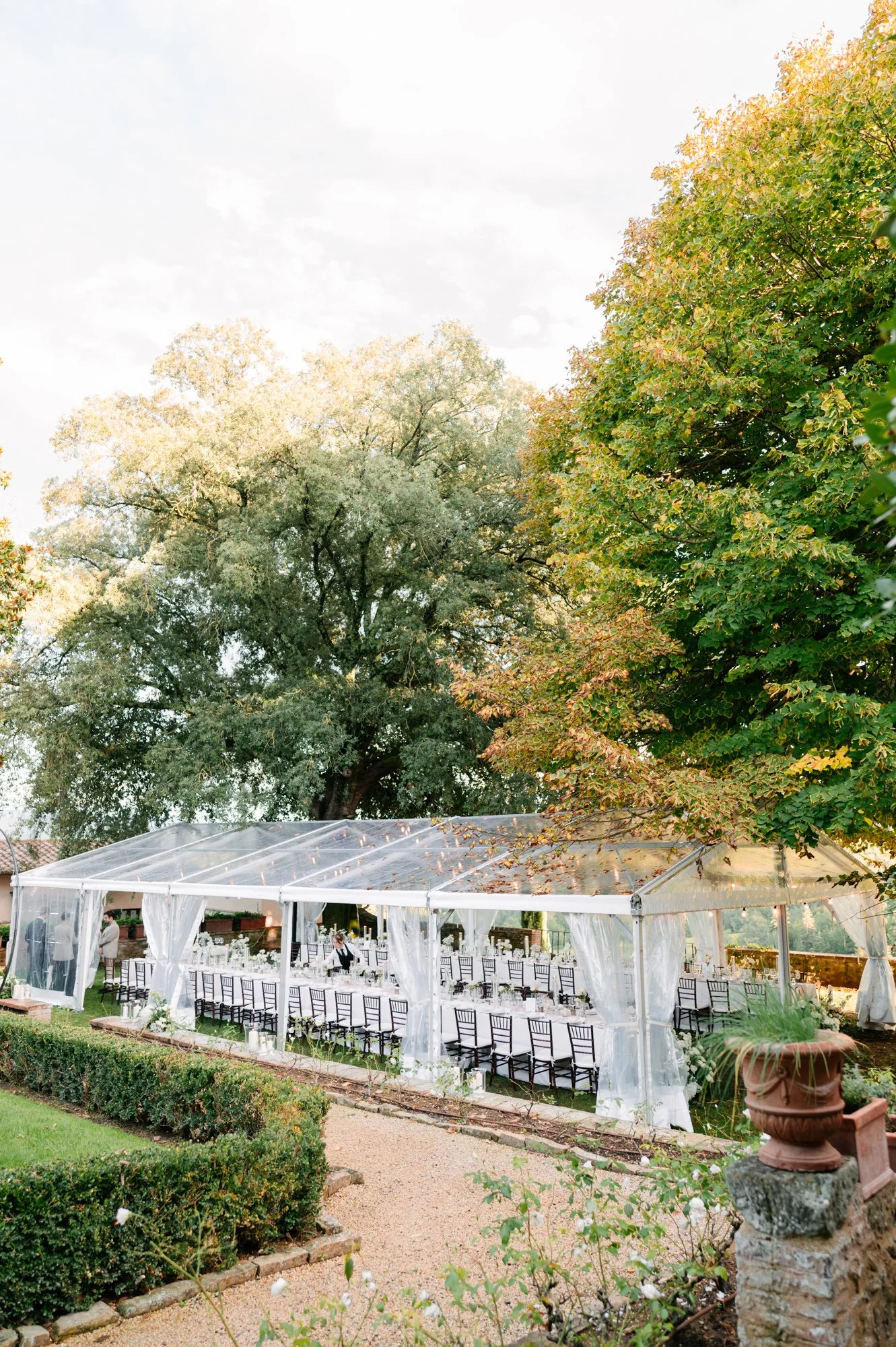 An outdoor wedding or event setup with a long table inside a clear tent, surrounded by greenery and trees, with chairs arranged along the table and decorative floral arrangements. Luxury editorial wedding photography in Tuscany. Destination wedding 