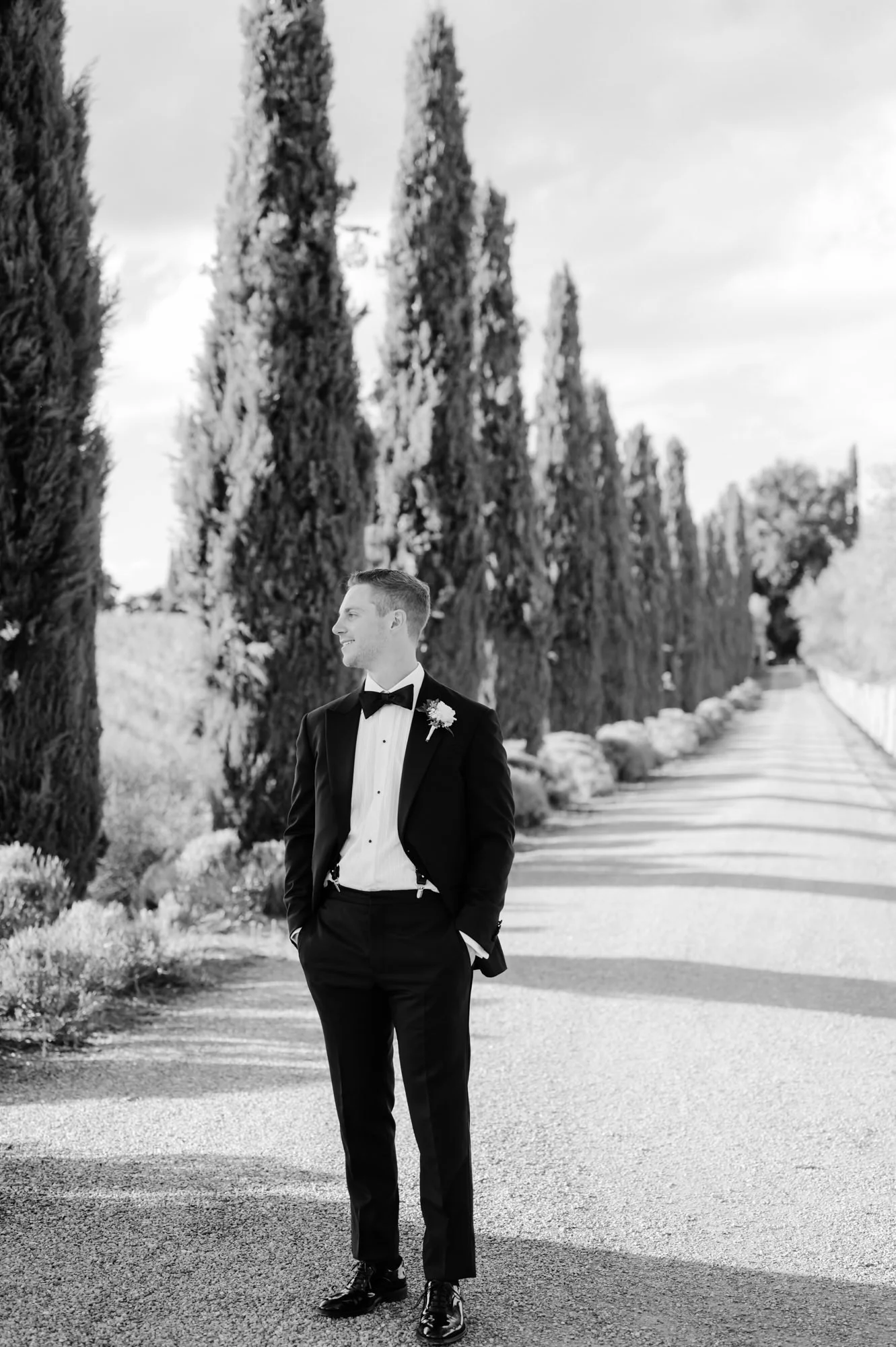 A man dressed in a tuxedo with a bow tie and boutonniere, standing outdoors on a tree-lined path, smiling and looking to the side in black and white.