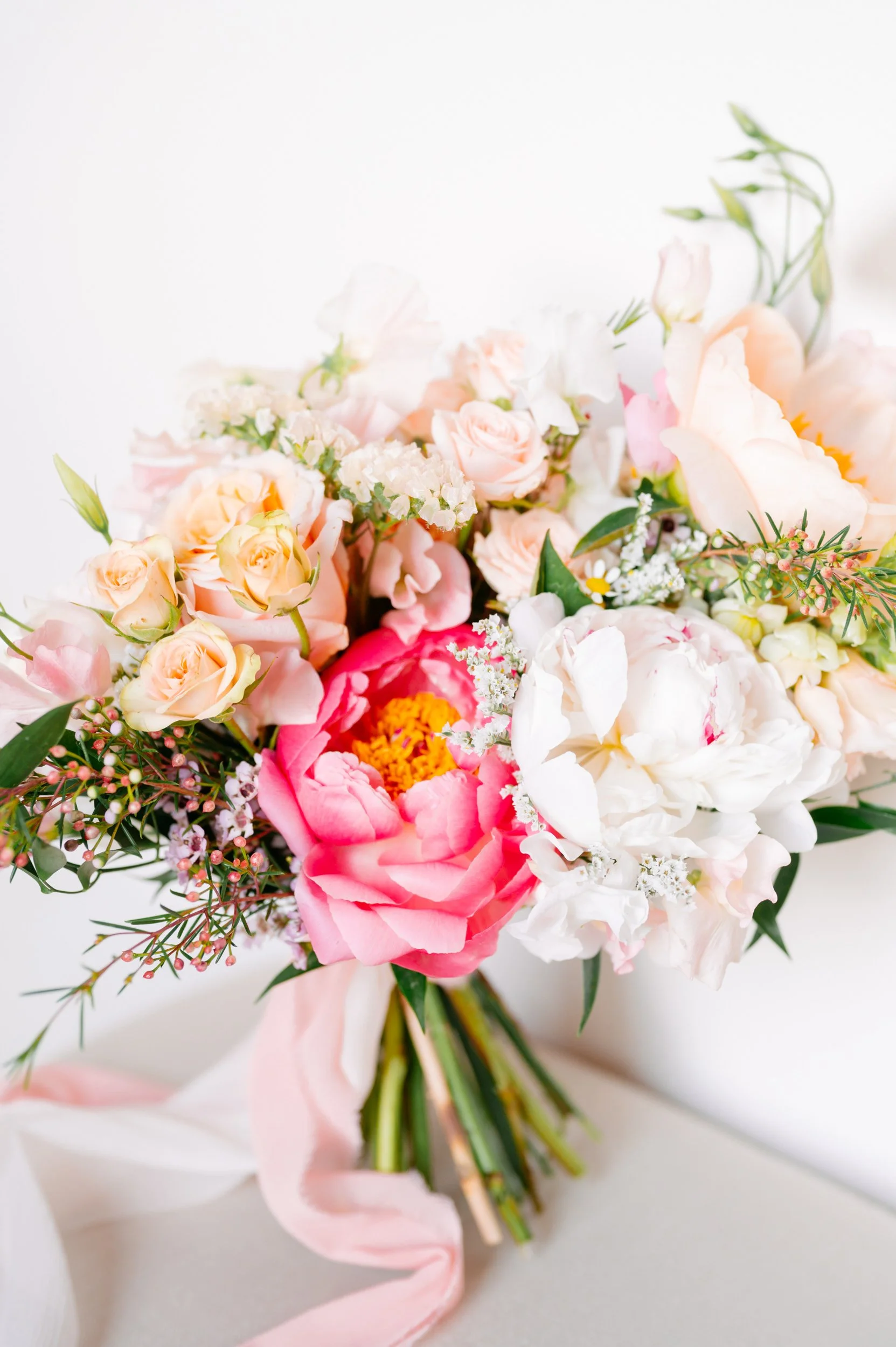 Colorful bouquet of pink, white, and peach flowers with greenery and pink ribbon on a white background.
