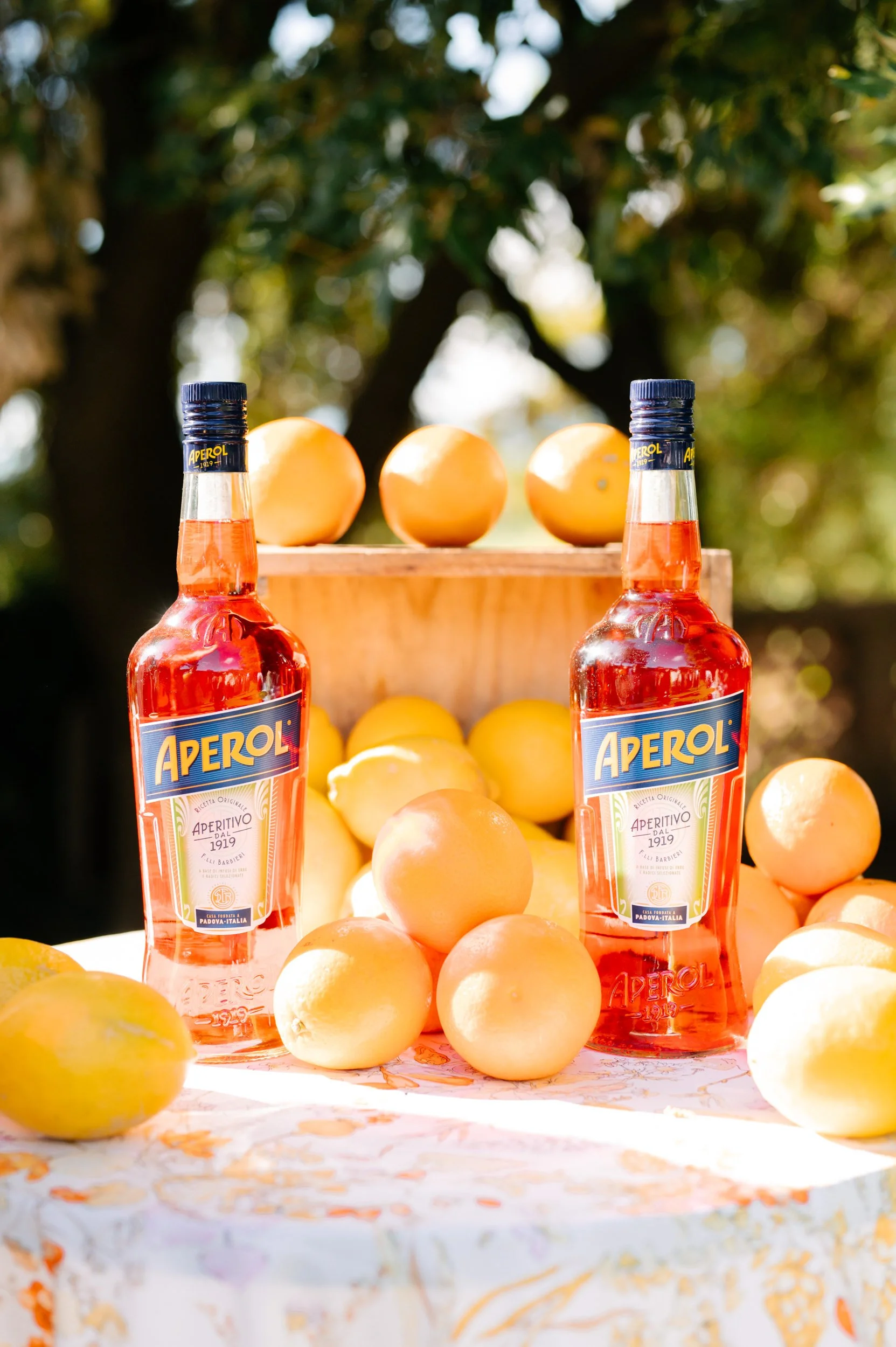 Two bottles of Aperol with oranges and lemons arranged outdoors on a table with a white cloth, against a background of green trees.