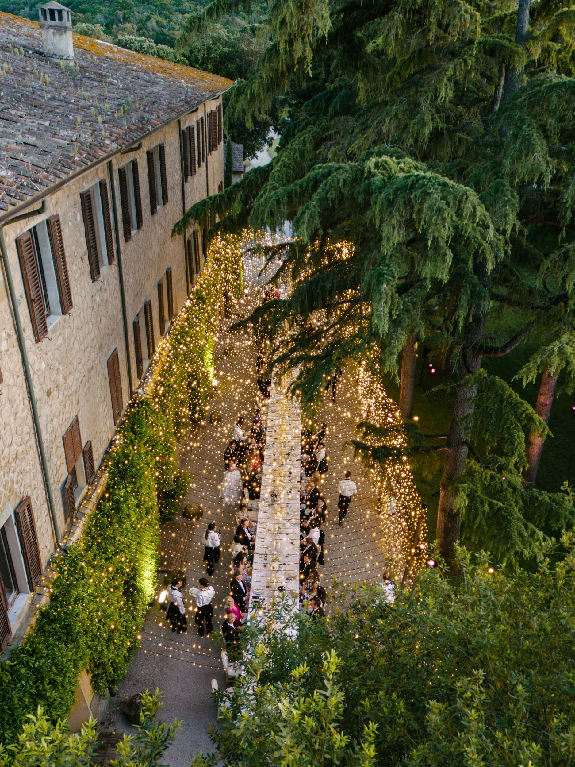 Outdoor wedding ceremony under string lights along a stone and brick building, with large trees nearby, during evening.