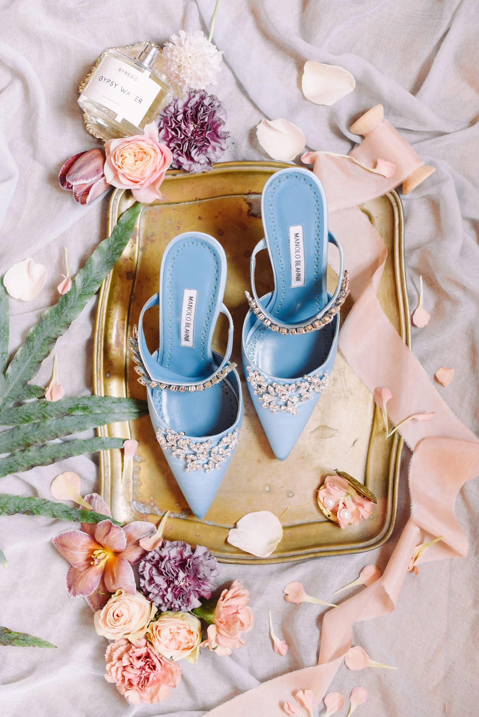 Pair of blue Manolo Blahnik heels decorated with rhinestones placed on a gold tray surrounded by pink and purple flowers, rose petals, a spray perfume bottle labeled 'Gypsy Water,' and a pink ribbon on beige fabric.