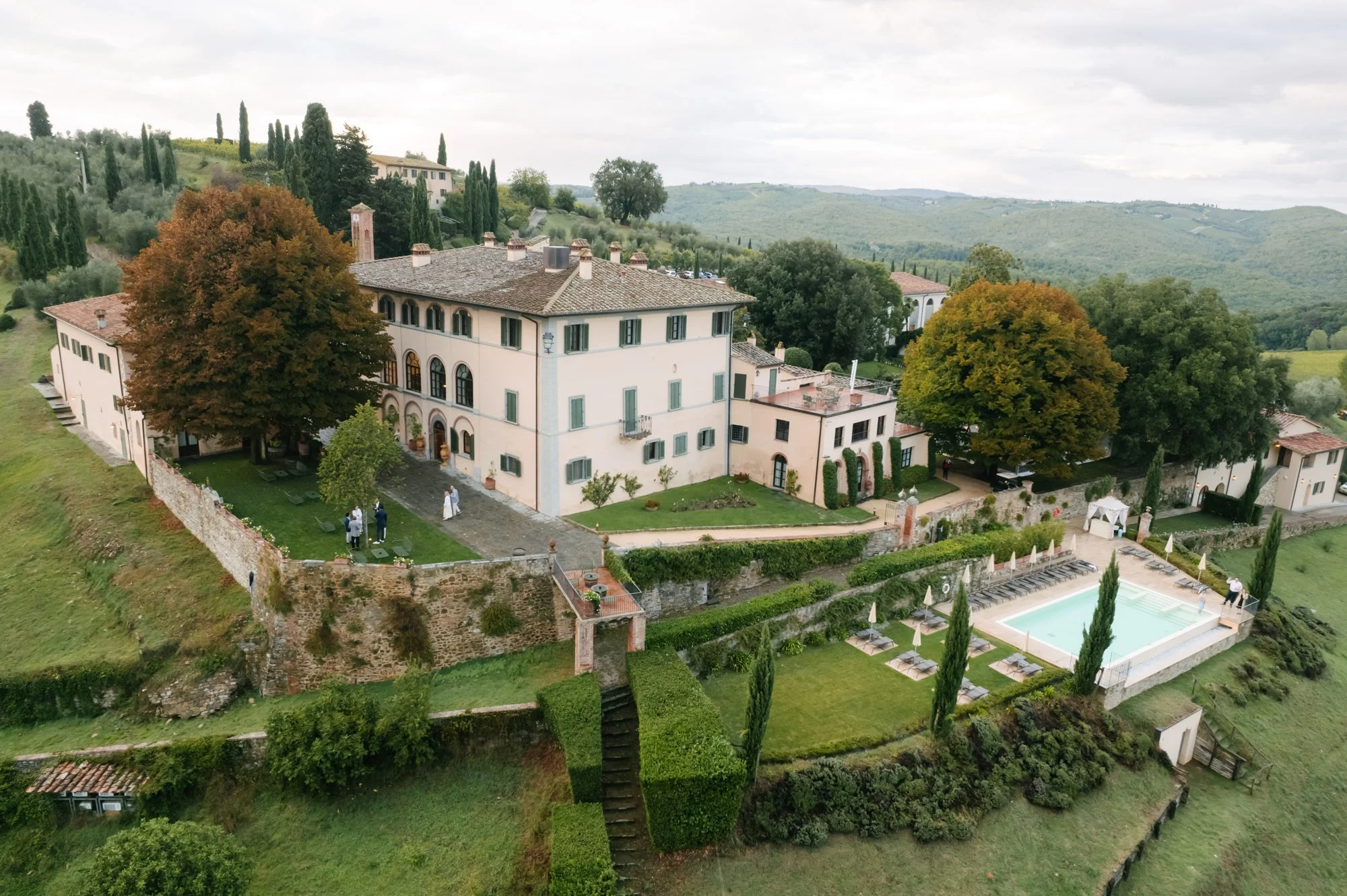 Aerial view of a large, historic hillside estate with a main building, a swimming pool, and lush greenery in the countryside of Tuscany, Italy.