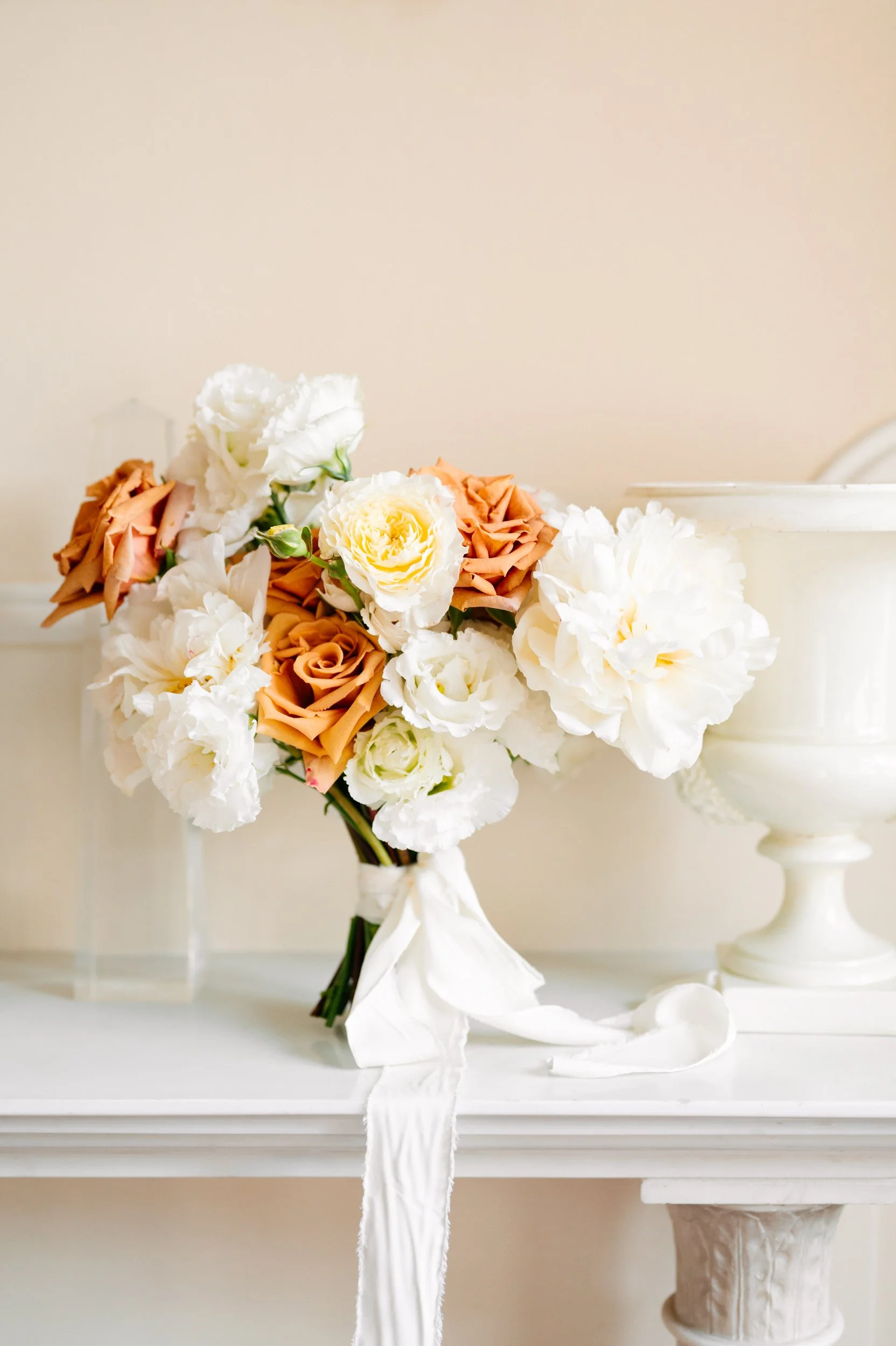A floral arrangement of peach roses and white peonies tied with a white ribbon, placed on a white mantelpiece.
