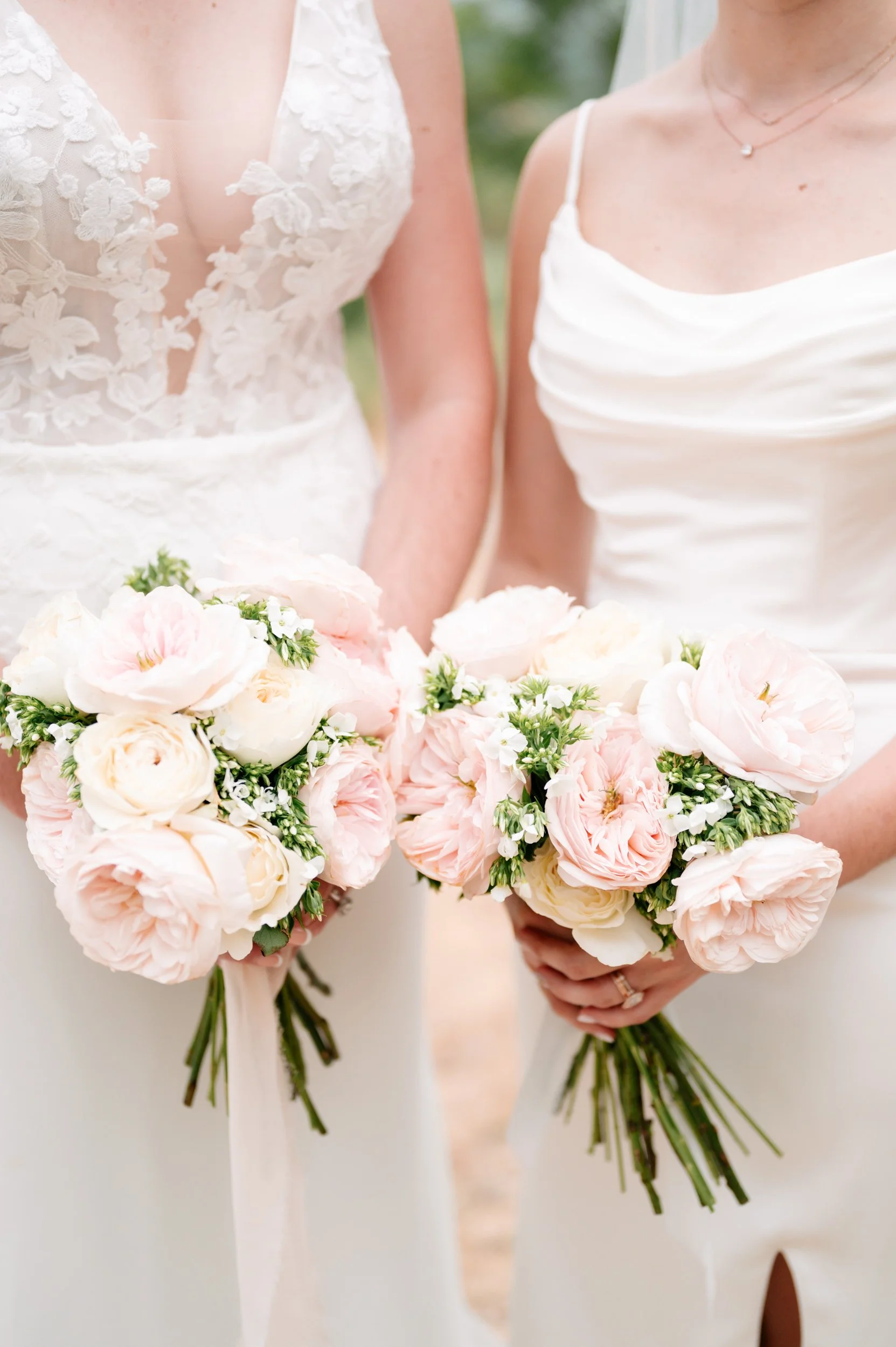 Two women in wedding dresses holding bouquets of light pink and white flowers.