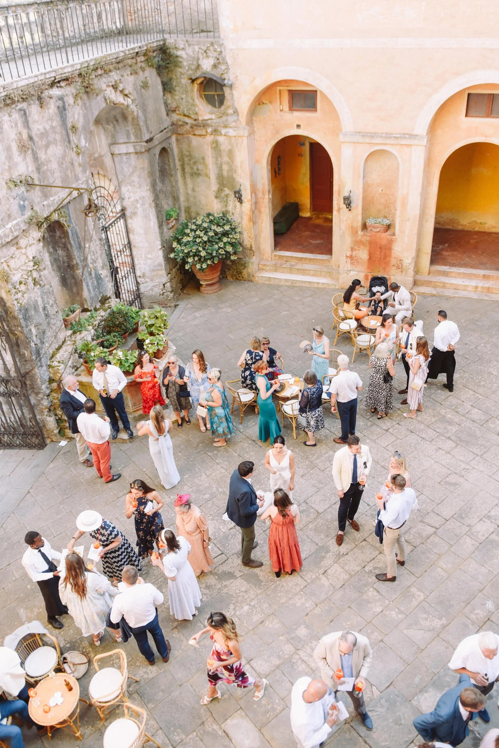 An outdoor courtyard filled with people attending a social gathering or party. Some are standing in groups, chatting, and holding drinks, while others are seated at tables. The setting features historic architecture with arched doorways, weathered wa