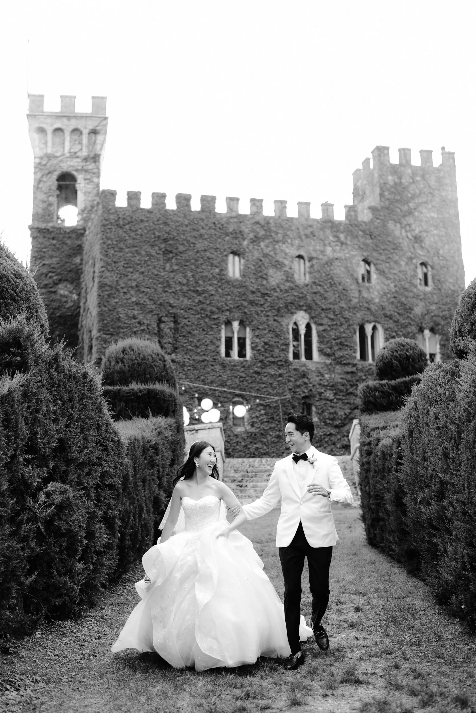 A bride and groom dancing on a lawn in front of a large, ivy-covered castle with turrets, surrounded by manicured bushes. The couple is smiling and holding hands, with the bride wearing a strapless wedding gown and the groom in a white tuxedo jacket 