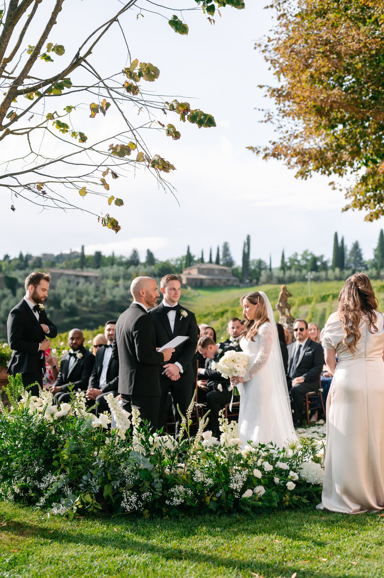 A wedding ceremony outdoors with a bride in a white gown holding a bouquet, a groom and officiant in suits, and guests seated on chairs. The setting features lush greenery and trees, with hills and a house in the background.
