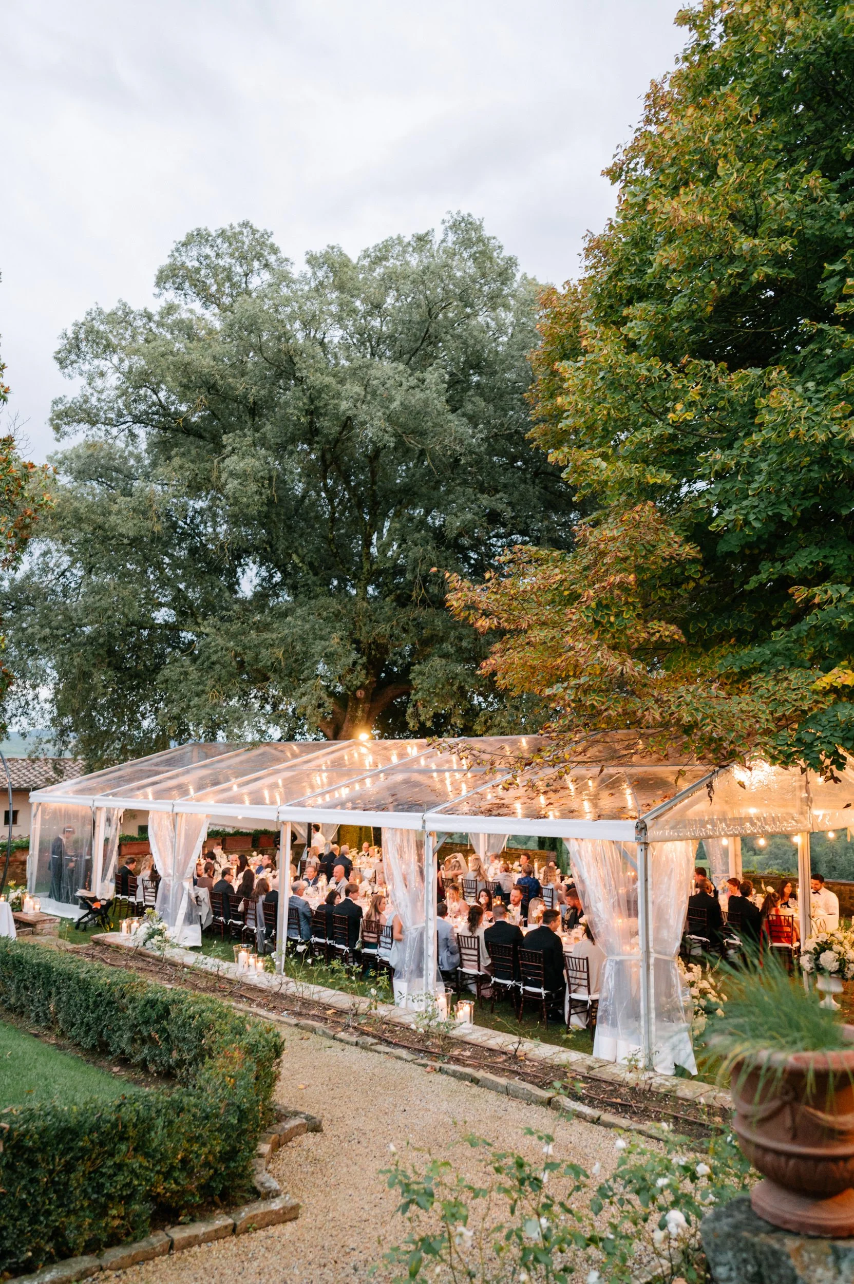 An outdoor wedding reception under a clear tent surrounded by trees, with numerous guests seated at decorated tables, under string lights in the evening.