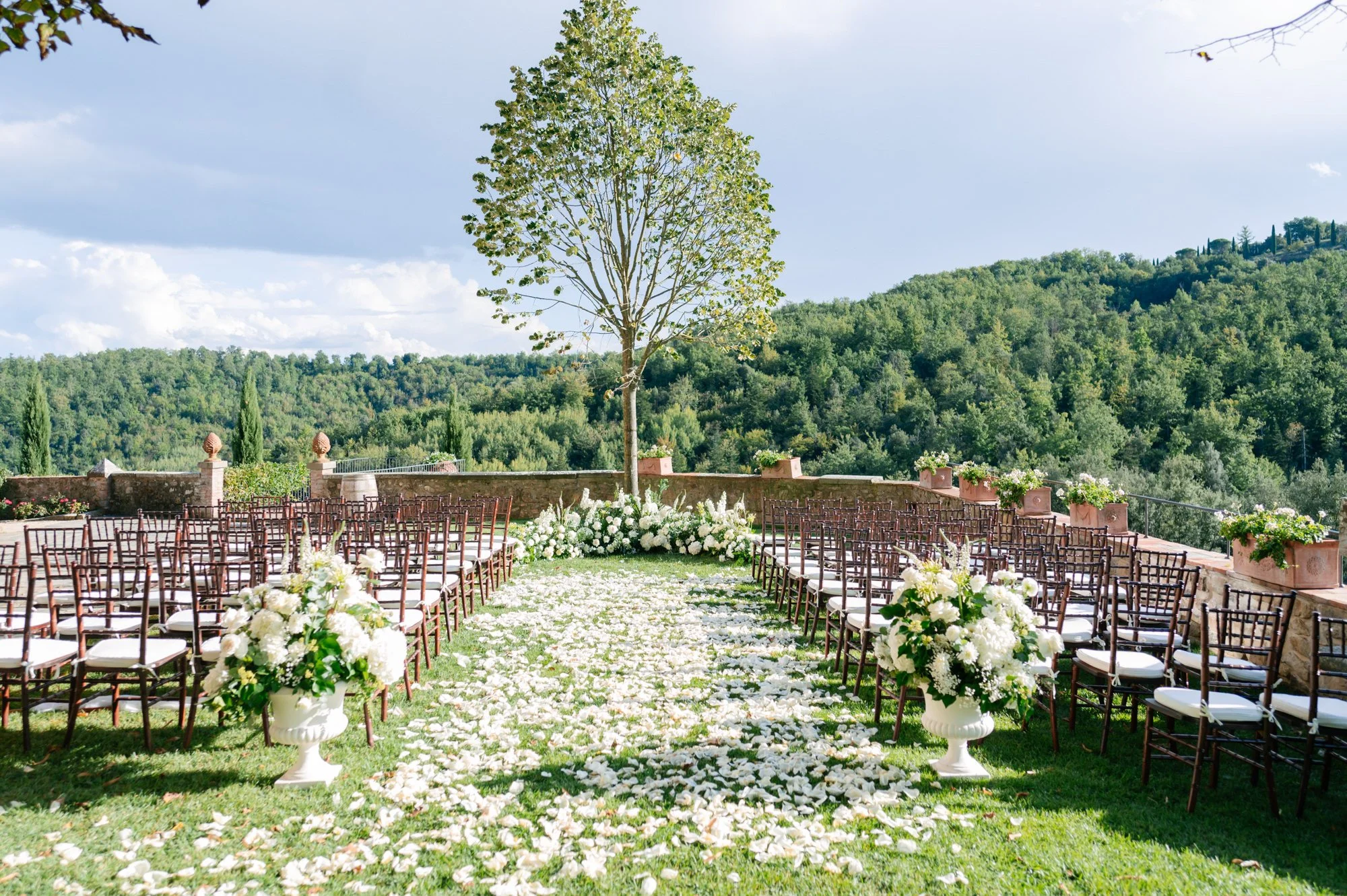 Outdoor wedding ceremony setup with rows of chairs, white flower arrangements, a central tree, and a scenic view of green hills and a cloudy sky in the background. Luxury editorial wedding photography in Tuscany. Destination wedding in Italy. 